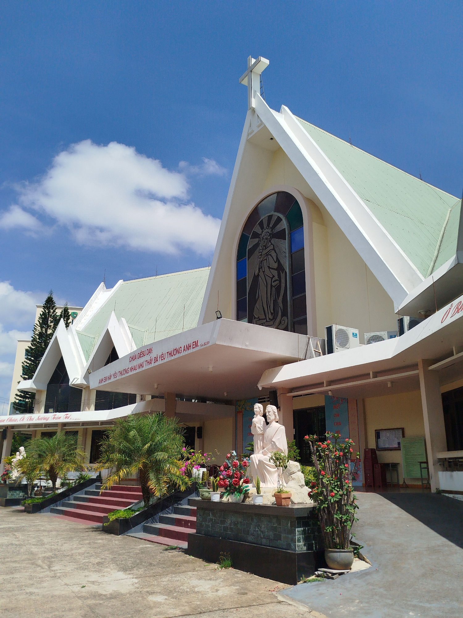 The photograph shows a church building, specifically the Giáo Xứ Thánh Tâm Pleiku (Sacred Heart Parish Pleiku), located in Pleiku Ward, Vietnam. The structure features a light yellow facade with white trim and a green-colored roof. Above the main entrance, there is a stained glass window with a depiction of a religious figure. A large white cross is positioned atop the highest point of the building.
In front of the church, there are decorative elements, including statues of religious figures and various potted plants and flowers. There is a raised concrete area with red steps leading to the main entrance. Two air conditioning units are mounted on the wall near the stained glass window.
The sky is blue with scattered white clouds, and the lighting indicates it is daytime. The overall scene suggests a well-maintained religious building in a tropical or subtropical climate.