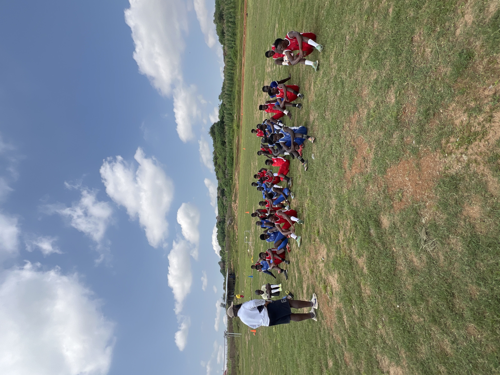 A group of young men in soccer uniforms are sitting on a grassy field, listening to a coach. The coach, wearing a white shirt and dark shorts, stands facing the group. The field appears to be part of a sports facility, with goalposts visible in the background. The sky is blue with scattered white clouds. The scene is set outdoors in a rural or semi-rural environment, possibly in or around Jos, Nigeria, given the red earth tones in the soil and the typical vegetation seen in such landscapes.