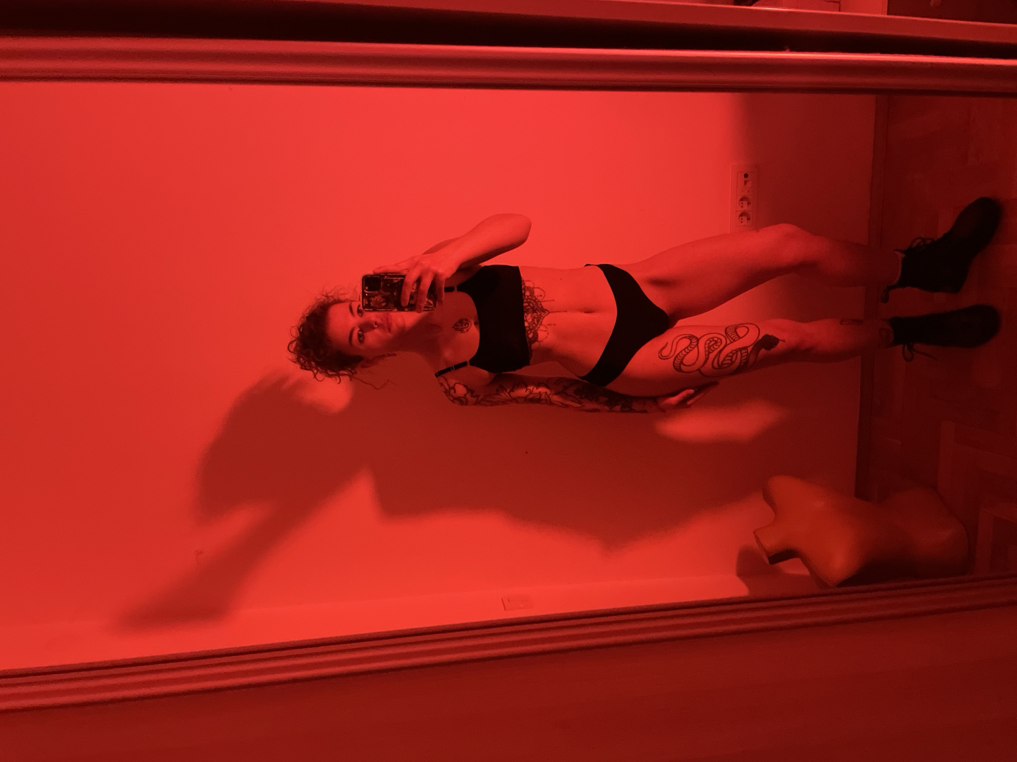 A woman stands in front of a mirror taking a selfie with her phone. She is wearing a black sports bra and black underwear. The environment has red lighting. The location is Boreti, Montenegro. There is a pillow on the floor. The woman has tattoos on her abdomen, thigh, and arms. She is wearing black boots. There is an electrical outlet on the wall.