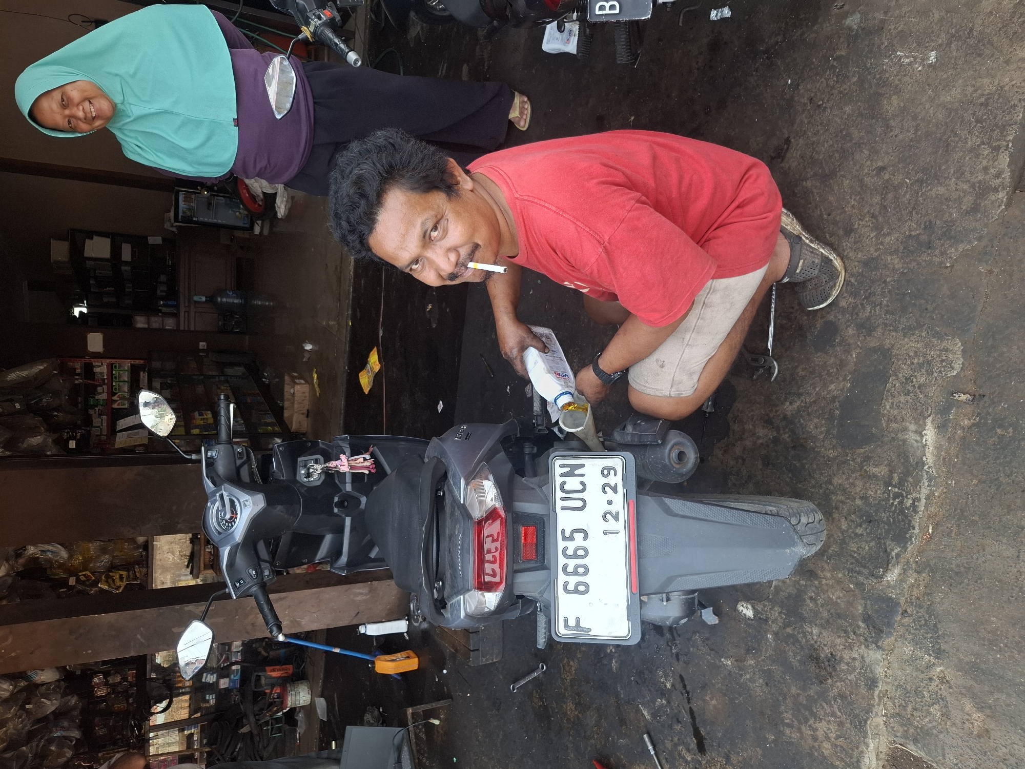 In Ciheulang Tonggoh, Indonesia, a man with dark, curly hair and a mustache, wearing a red t-shirt and light-colored shorts, crouches next to a grey scooter. He is smoking a cigarette and appears to be pouring liquid from a white bottle into the scooter's engine, using a funnel. The scooter has a license plate reading "F 6665 UCN" with "12.29" below it. A woman wearing a teal hijab and purple shirt stands behind him, smiling. The scene takes place in what appears to be a workshop or repair shop, with shelves of parts and tools visible in the background. The ground is dirty and wet, with oil stains.