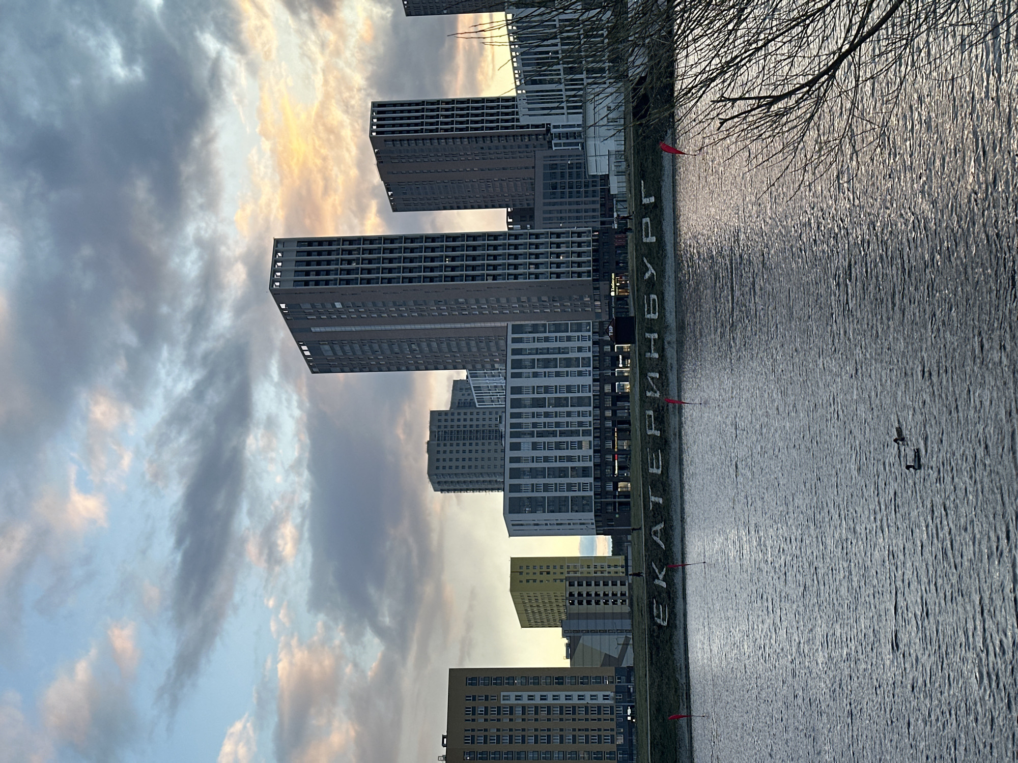 The image captures a cityscape with a focus on modern high-rise buildings reflected in a body of water, likely a river or lake, during sunset. The sky features a mix of gray clouds and golden hues from the setting sun. Along the water's edge, large Cyrillic letters spell out "ЕКАТЕРИНБУРГ" (Yekaterinburg) in Russian. Small red flags are interspersed with the lettering. Bare tree branches are visible on the right side of the frame, closer to the viewer. The water's surface is textured by wind or current. Two swimming birds are visible on the right. The architecture is contemporary, with numerous apartment buildings of varying heights and colors. The location is Yekaterinburg, Russia.