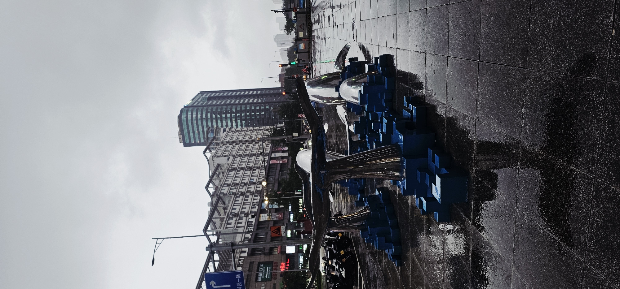 This image depicts an urban street scene in Jiulongpo District, China, under an overcast sky. The foreground features dark, square-tiled pavement, visibly wet and reflective from recent precipitation. Several metallic sculptures are positioned centrally, resembling abstract whale tails or streamlined forms, supported by blue, pixelated, cube-shaped bases of varying heights. The sculptures exhibit high reflectivity, mirroring the surroundings.
In the midground, a street runs horizontally. To the left, a row of motorcycles is parked on the wet ground. A tall, dark streetlamp stands prominently on the left. Various street signs with Chinese characters are visible, including a blue directional sign with a right-pointing arrow and the text "科园一路" (Keyuan 1st Road). Traffic lights are visible further down the street on the right, displaying a green light. Indistinct vehicles and a few distant human figures are present on the street.
The background is dominated by city buildings. On the left, a multi-story building with a light-colored facade, numerous windows, and visible external air conditioning units exhibits a tiered roofline structure. To its right, a significantly taller, modern high-rise building with a dark, possibly glass, facade and a curved profile rises against the sky. The sky is uniformly overcast and grey, providing diffused lighting to the scene. Distant urban structures are faintly visible on the horizon to the far right.