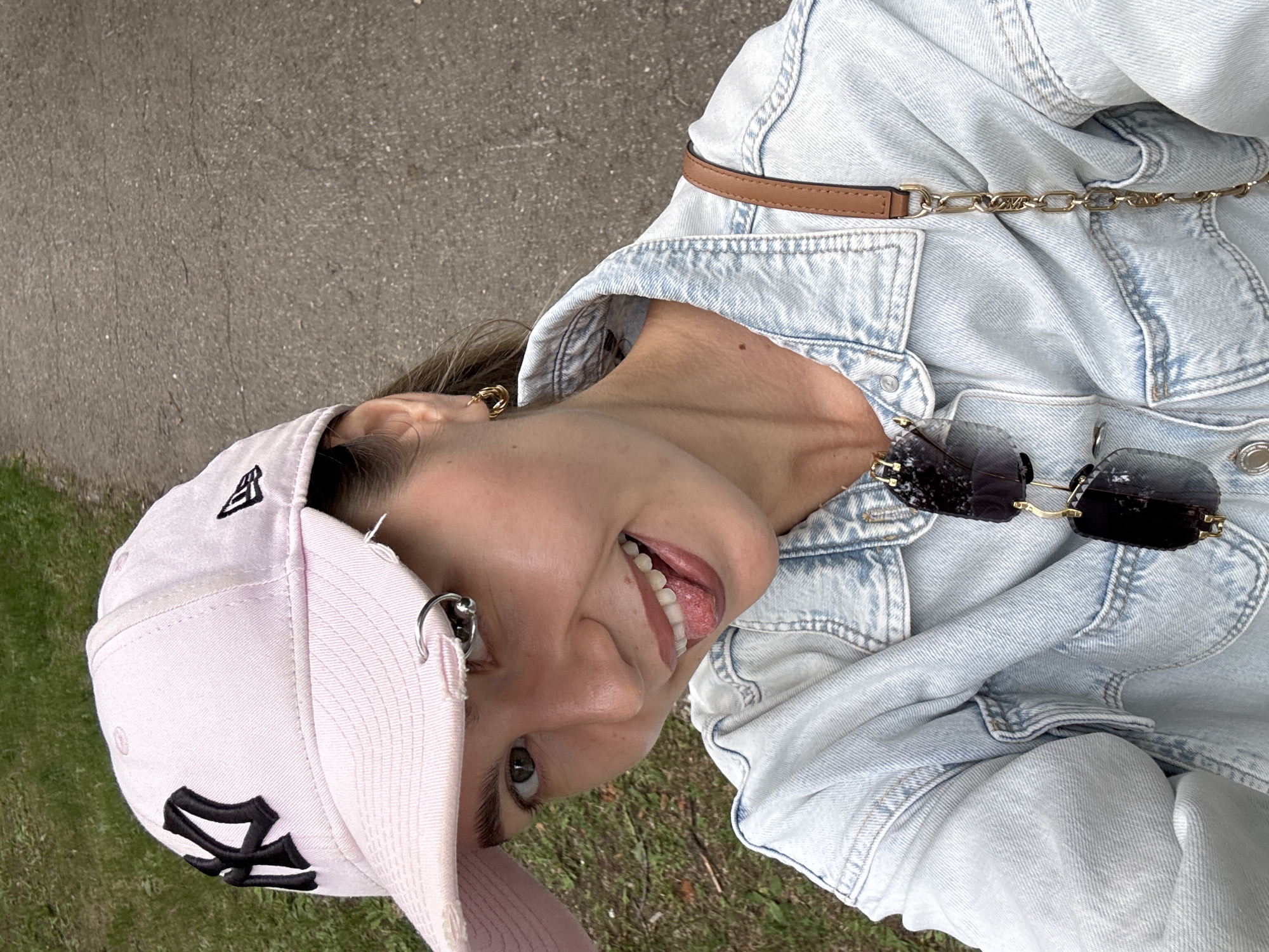 A young woman is taking a selfie outdoors on a paved path. She is wearing a light pink baseball cap with a black New York Yankees logo and a New Era logo. The cap has a distressed look with a hole in the brim and a silver hoop earring through it. She has on a light blue denim jacket with a collar and chest pockets. A brown strap with a gold chain is visible over her shoulder. She is smiling and sticking her tongue out slightly. Dark sunglasses are hanging from the neckline of her jacket. The background shows green grass and the gray paved path.