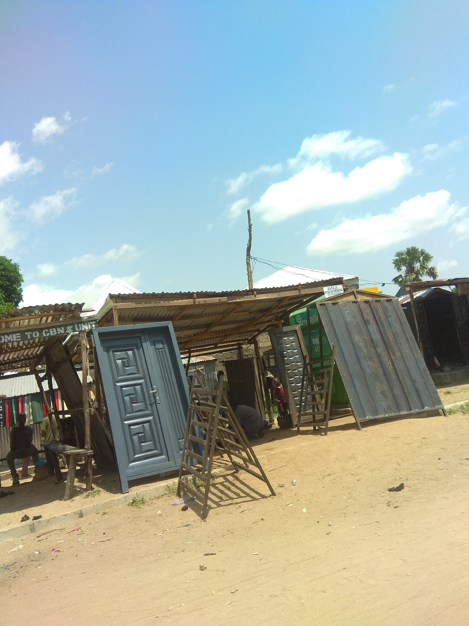 This media file depicts an outdoor market scene in Jalingo, Nigeria, under a bright, partly cloudy sky, suggesting a mid-morning or afternoon timeframe. The setting is dusty and arid, typical of many Nigerian landscapes.
The primary subjects are various doors displayed for sale in front of a makeshift stall. A large, dark gray double door with an intricate geometric pattern is prominently featured, leaning against a wooden structure. To its right, a tall, flat, dark metal sheet leans against a green container. Behind these, more doors and metal sheets are visible, along with wooden ladders and scaffolding, suggesting ongoing construction or sales activity.
In the background, a corrugated metal roof shelters the stall, supported by rough wooden beams. A white-tiled roof of a building is visible further back, along with a palm tree. The scene is sparsely populated. Two individuals are partially visible near the stall: one sitting on a wooden bench, appearing to be a vendor or customer, and another person partially obscured behind the display.
There are several instances of visible text. On a sign above the stall, the words "COME TO CBN UNIT" are clearly legible. On the green container, the word "mobile" and the number "4G" are visible, indicating a telecommunications presence or service. Another sign with text is partially visible in the upper right portion of the image.
The overall impression is one of commerce in a developing area, with goods displayed outdoors and the elements playing a significant role in the environment. The bright sky and scattered clouds suggest pleasant weather.