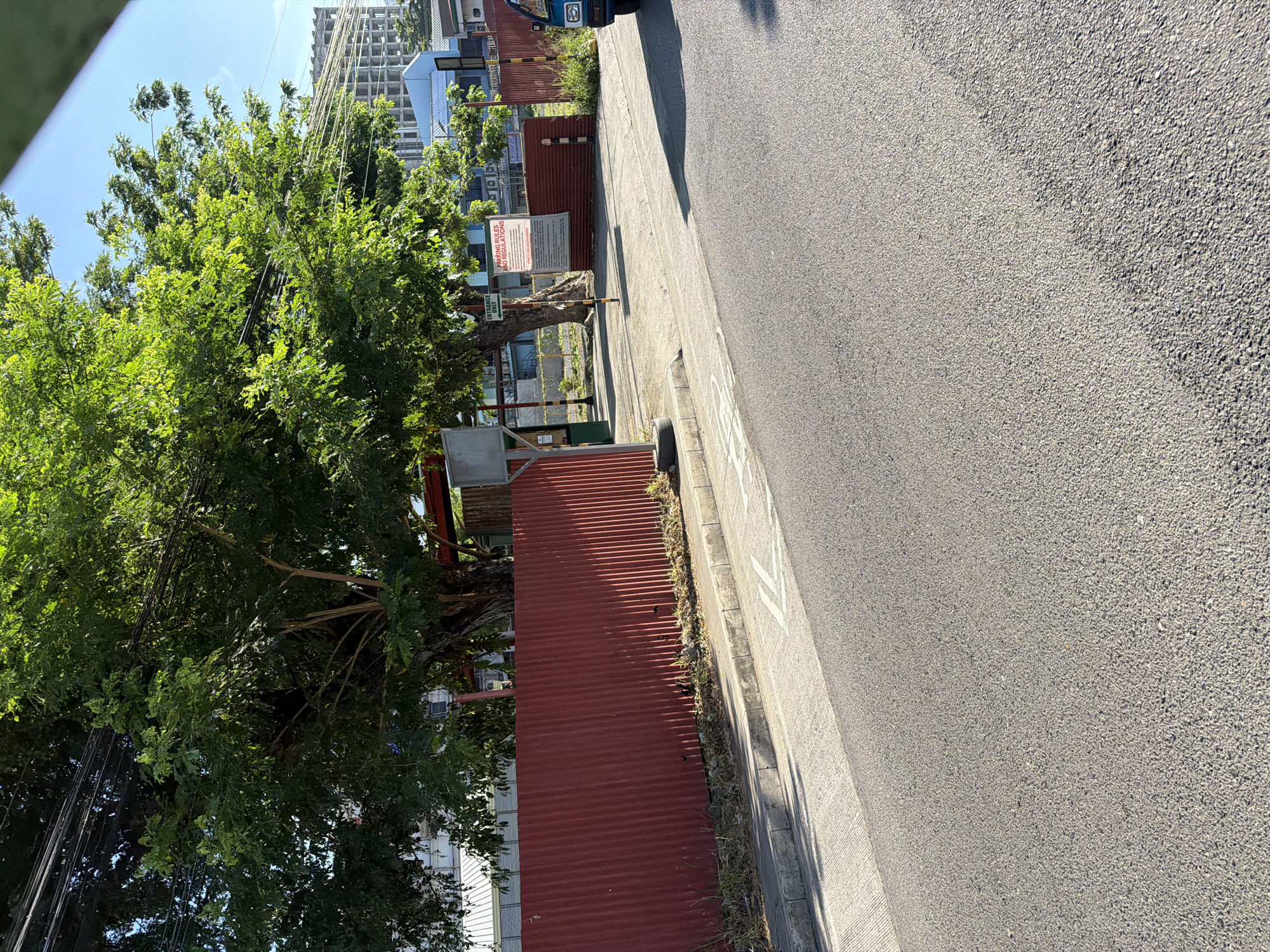 In Davao City, Philippines, a wide asphalt road runs parallel to a corrugated red metal fence. Behind the fence, a large, leafy tree with visible power lines overhead casts shadows on the paved ground. Beyond the tree, a building under construction, with visible concrete pillars and floors, dominates the background. A blue and white building is also visible. Signs are posted near the entrance to the property, with one prominently displaying "PARKING RULES AND REGULATIONS." A blue vehicle is partially visible on the right side of the image. The sky is clear and blue.
