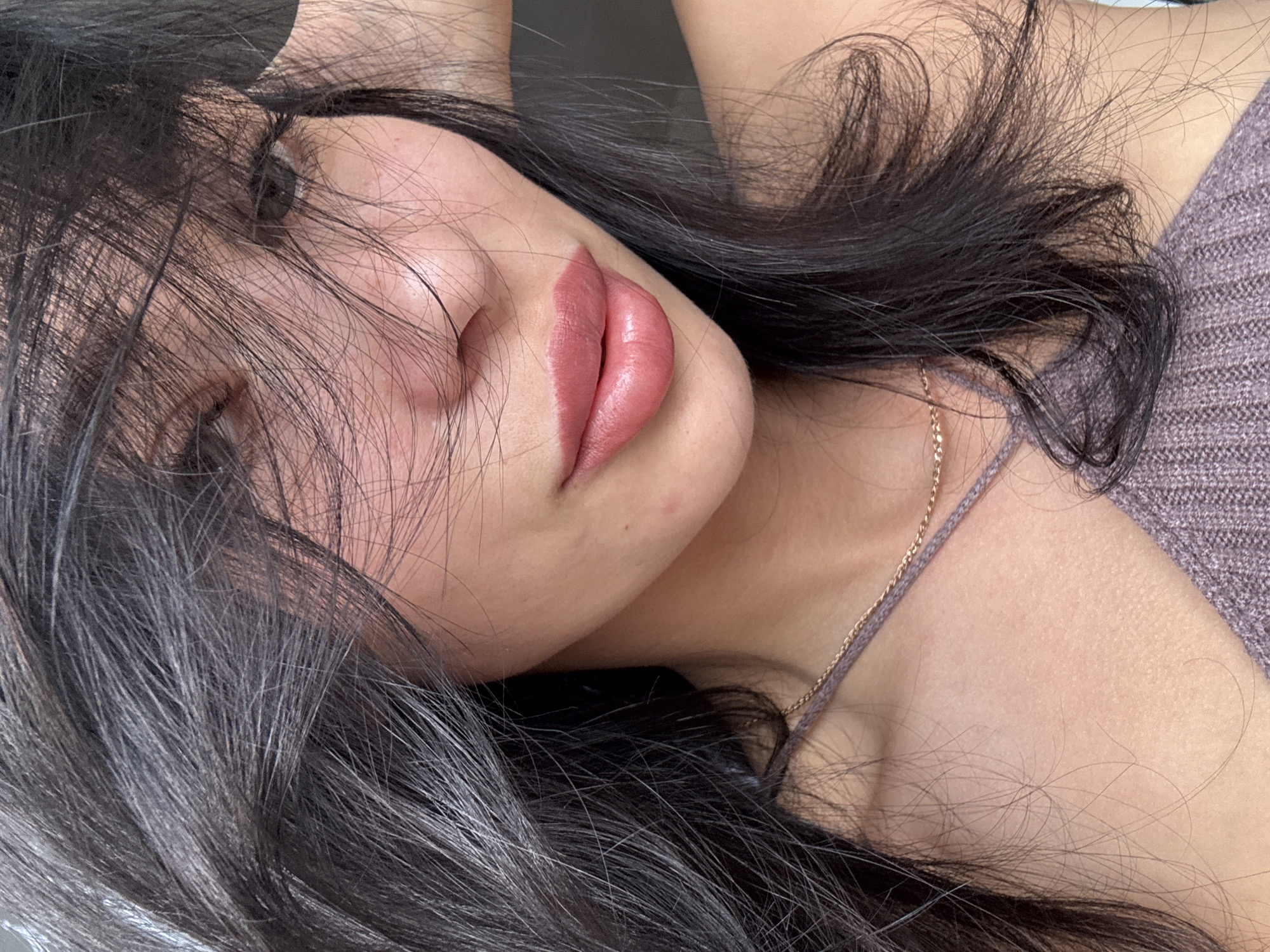 The image is a close-up, overhead shot of a young woman lying down. She has long, dark, slightly messy hair that falls across her face and shoulders, obscuring most of her features. Her eyes are closed, and her face is turned to the side, with her lips slightly parted and painted a rosy pink. A delicate gold chain rests on her collarbone, just visible beneath the collar of her purple, textured top. The lighting appears soft and diffused, suggesting an indoor setting, possibly a bedroom. The overall mood is intimate and relaxed, with a hint of vulnerability. There is no visible text in the image. The location context of Chaikovsky, Russia, does not provide any specific visual cues within the image itself.