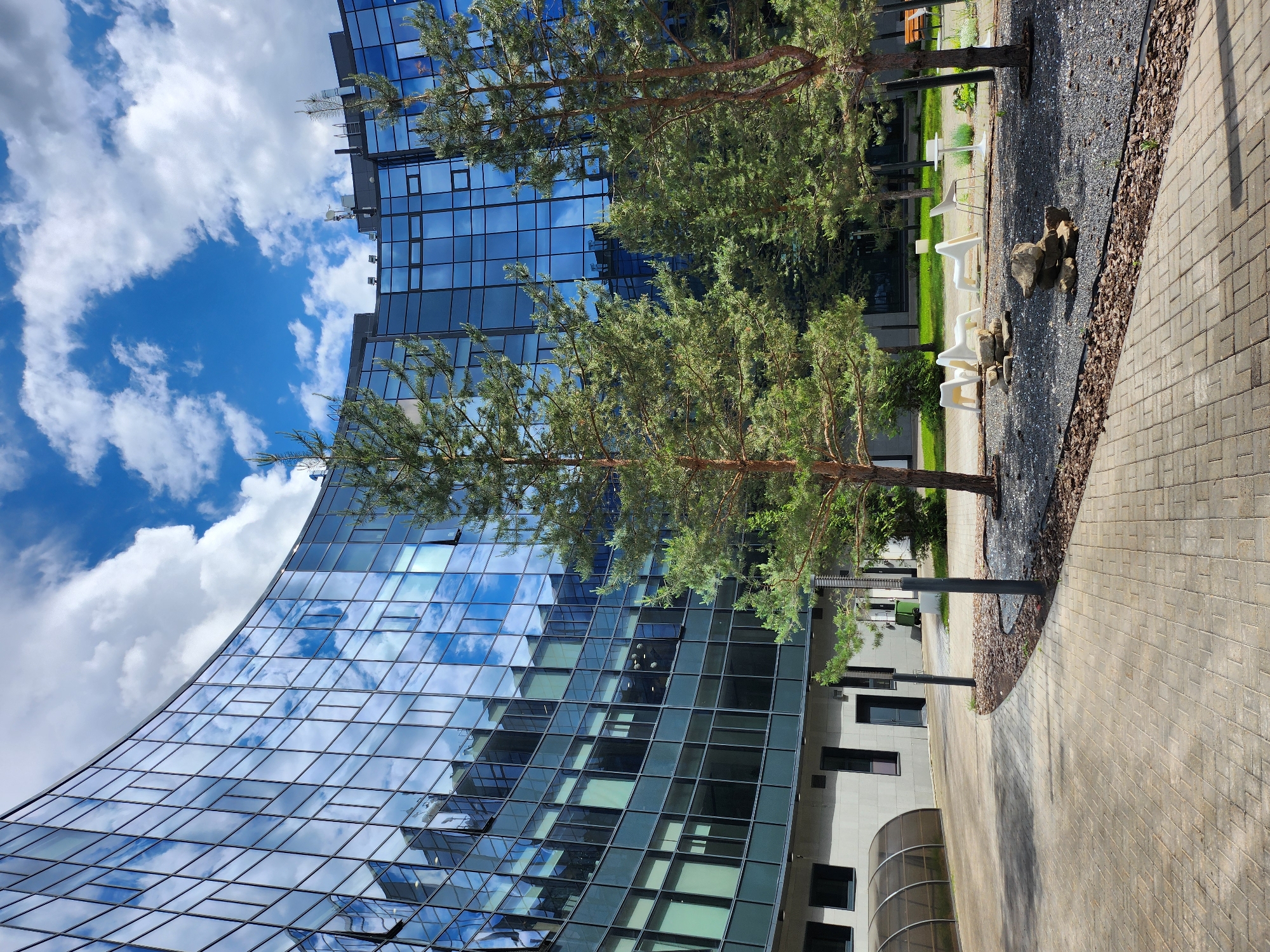The image presents an outdoor scene featuring a modern building with a curved, glass-clad facade reflecting the sky and clouds. Two trees with sparse, green foliage are positioned in front of the building, partially obscuring the view. A paved walkway runs along the side of the building, adjacent to a patch of dark gravel containing a collection of rocks. Some white chairs are visible in the background. The location appears to be an urban or suburban setting, specifically in the city of Unknown, Unknown. The light suggests it is daytime.