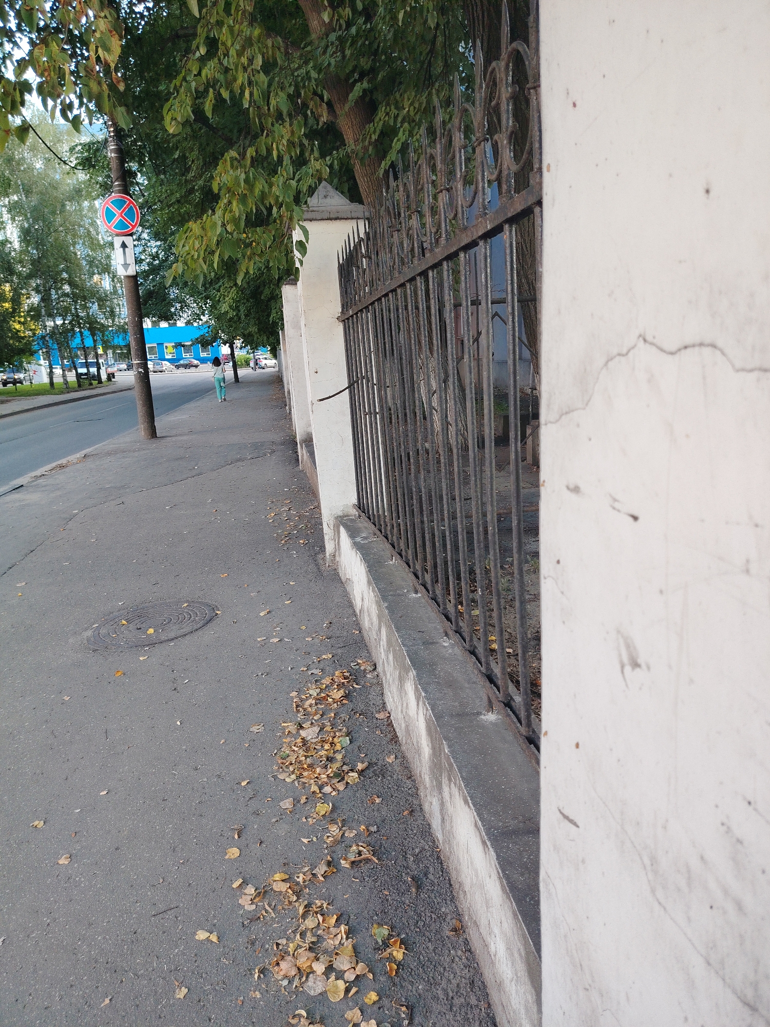 A full shot captures a sidewalk bordering a decorative wrought-iron fence with ornate scrollwork. The fence is set against a textured, light-colored wall. Fallen leaves litter the pavement and the base of the fence. To the left, a street is visible with cars and a building with a blue facade in the distance. A young person wearing a light blue outfit walks away from the camera on the sidewalk. A traffic sign, circular with a red border and a blue cross, is mounted on a pole near the street. The scene suggests a city environment, possibly in Nizhny Novgorod, Russia, given the context provided.