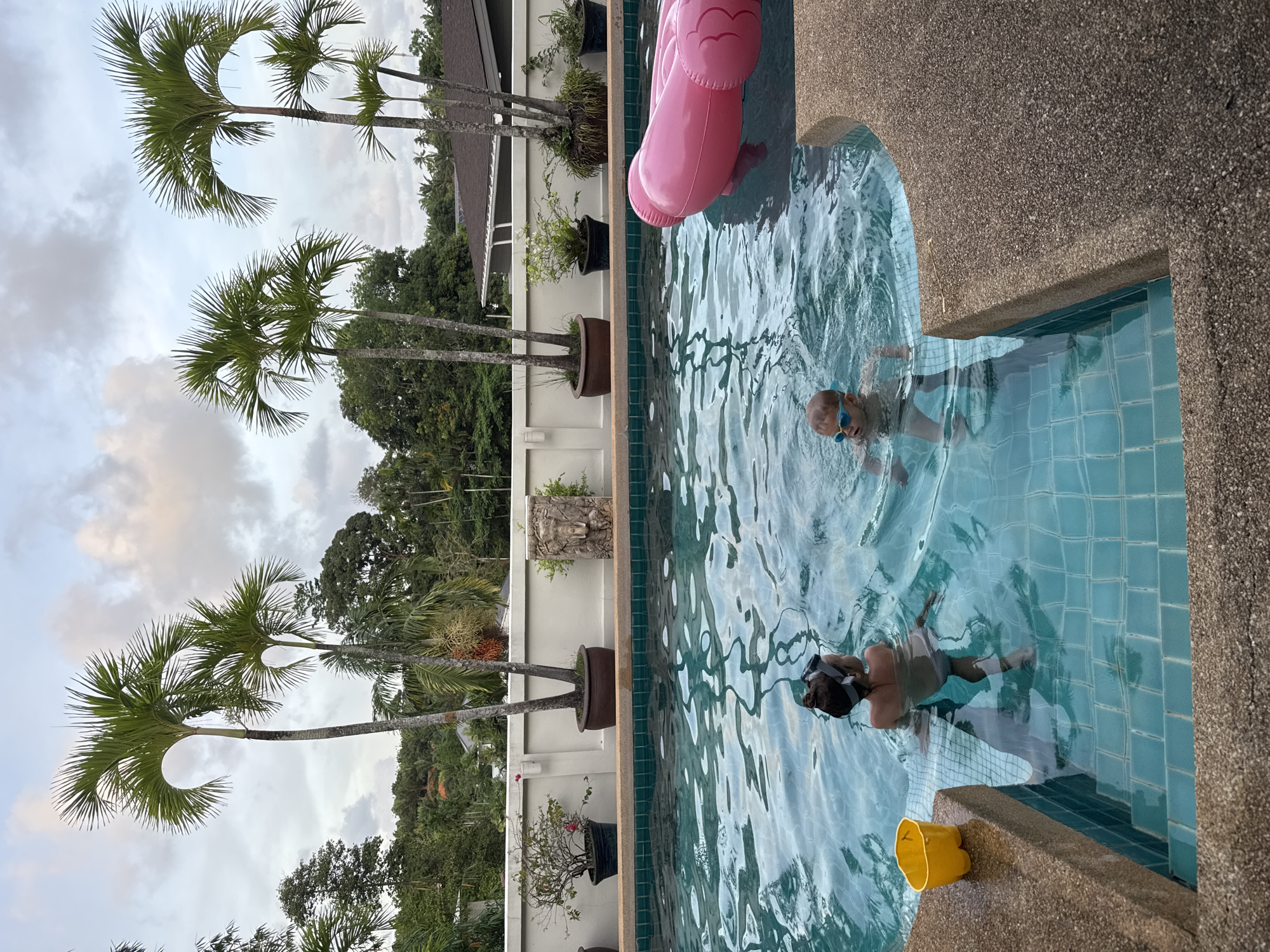 The scene captures two people swimming in a pool in Rawai, Thailand. One person wears swimming goggles and appears to be a child. A pink flamingo float is visible in the pool. A yellow bucket sits at the pool's edge. The pool is surrounded by a decorative white wall adorned with potted plants. Tall palm trees line the edge of the wall, with a dense green forest visible in the background under a cloudy sky. The photo appears to be taken from an elevated perspective, looking down into the pool area.