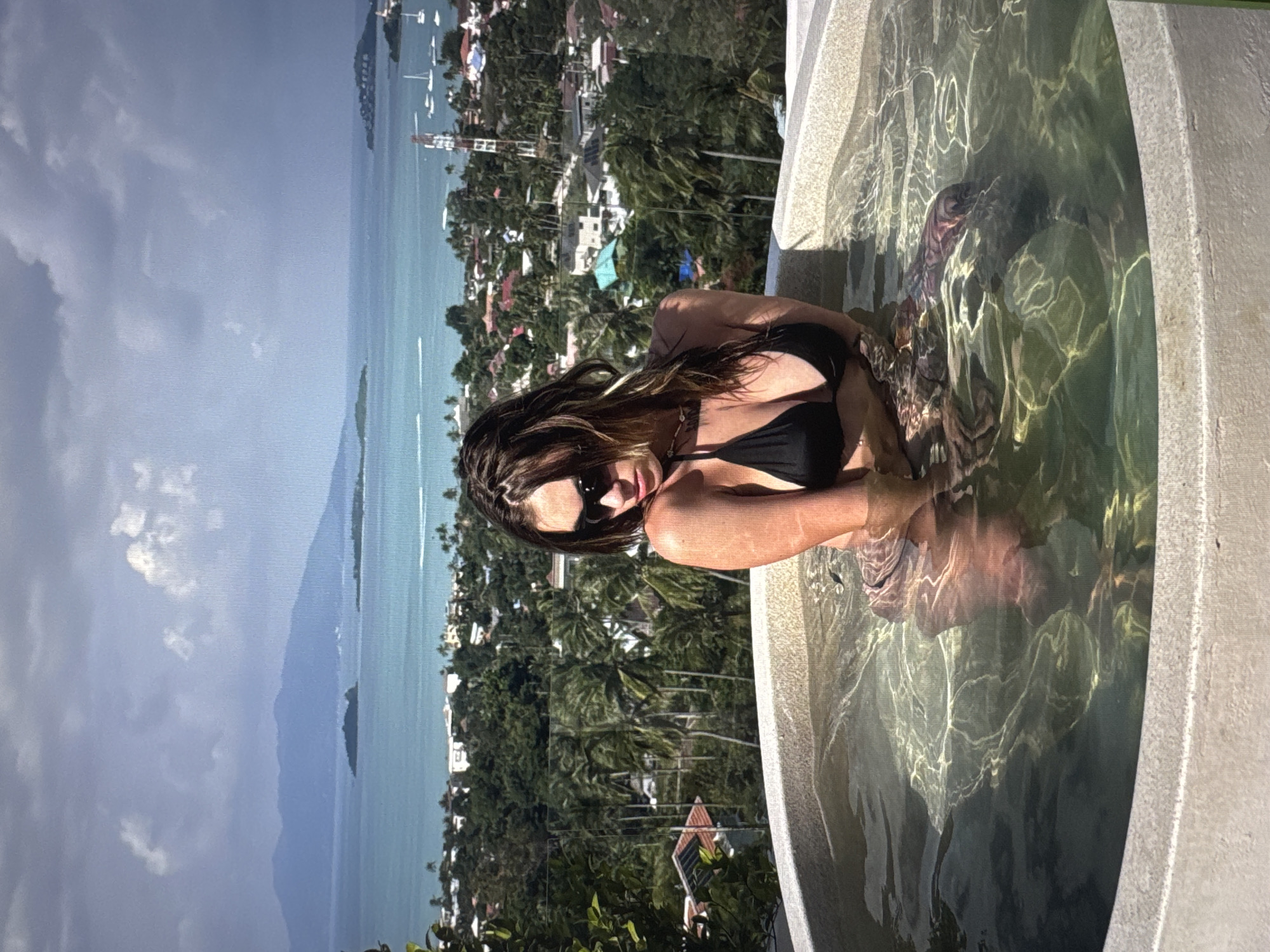 A woman in a black bikini is sitting in a circular hot tub with a view of islands and a town. She is wearing sunglasses and has her hair down. The water in the hot tub is clear and reflects the sunlight. The background shows a body of water with several islands and a town nestled among palm trees. The sky is partly cloudy. The hot tub appears to be on a balcony or terrace. This image was taken in Moscow, Russia.