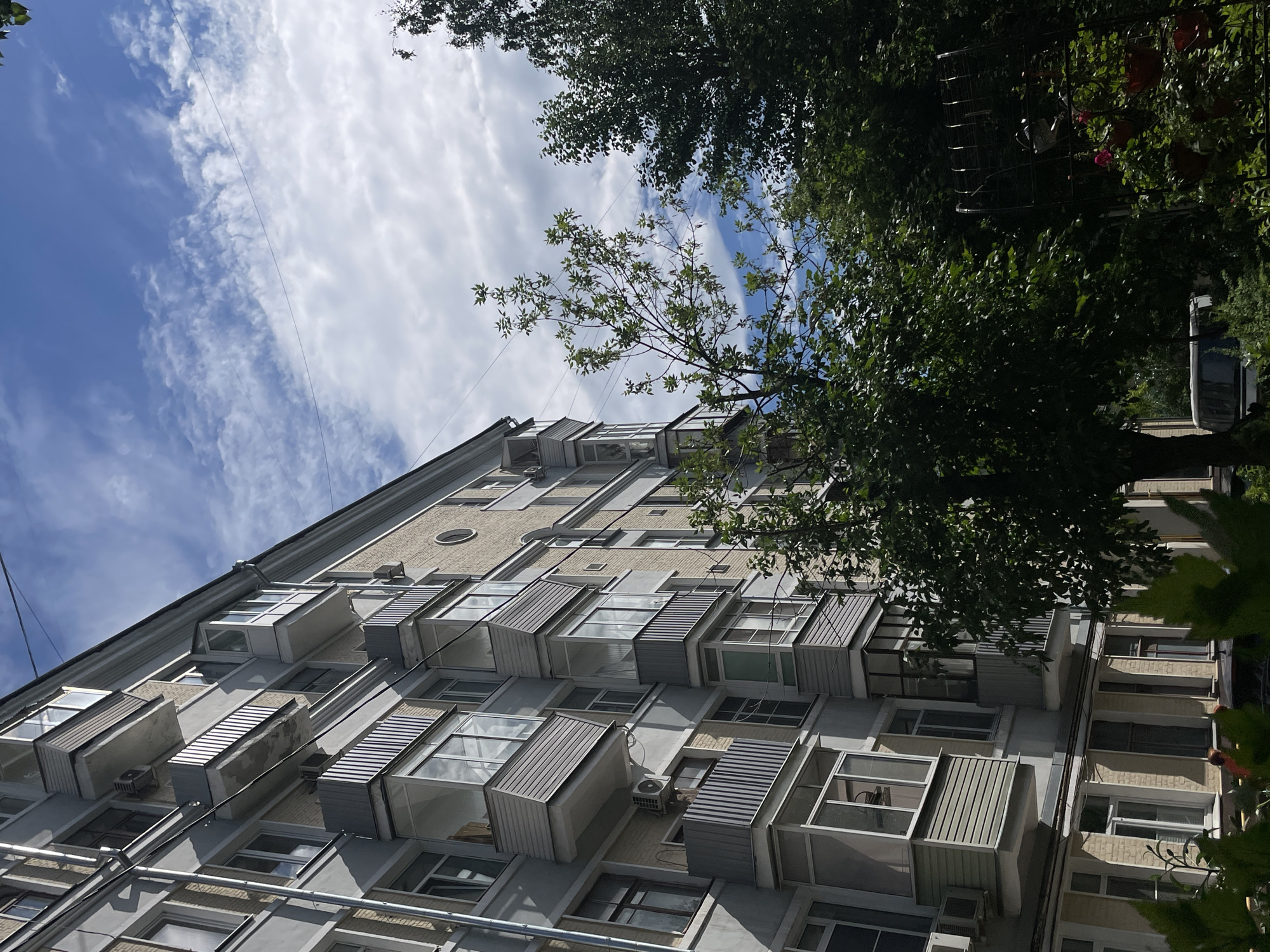 The image shows the facade of a multi-story apartment building, angled upwards towards a bright blue sky with scattered white clouds. Lush green trees frame the top and right side of the image, partially obscuring the building. The building itself is light gray with numerous balconies, many of which are enclosed with glass and have metal or wooden slatted coverings. Some balconies have air conditioning units mounted below them. The architecture suggests a Soviet-era or post-Soviet construction, common in Moscow. The building's facade features light brown brick accents around some windows. The overall scene evokes a bright, sunny day. The angle of the shot and the presence of trees indicate an outdoor perspective, likely from the ground or a lower floor of an adjacent building. No people are visible, and the focus is on the architecture and the natural elements. The time of day appears to be daytime, given the bright sky and shadows. There is no visible text in the image.