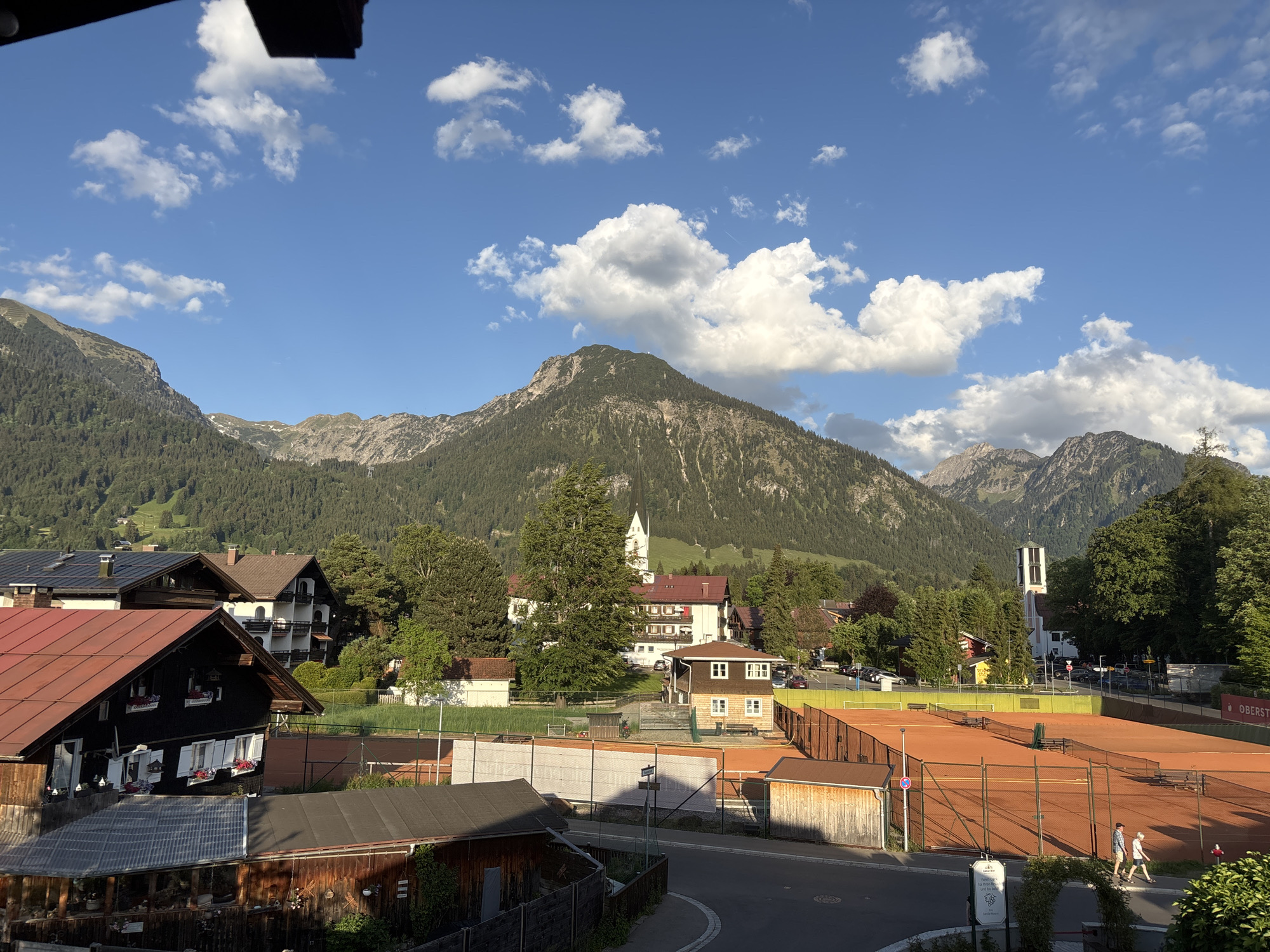 This image shows a view of Oberstdorf, Germany. In the foreground are outdoor tennis courts, with two people walking alongside them. Above, there is a mix of residential buildings, trees, and greenery, scattered throughout the town. In the distance, forested mountains rise against a clear blue sky with scattered white clouds. The architecture suggests a traditional Alpine style. The lighting suggests a sunny day.