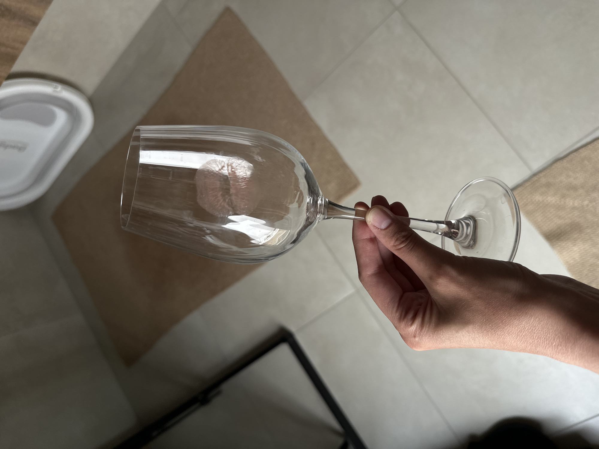 The image shows a hand holding a clear wine glass. Inside the glass, there is a crumpled light pink object, possibly a napkin or tissue. The background consists of light-colored tiled flooring with beige rugs and a glimpse of what appears to be furniture with black legs. The image does not offer sufficient information to determine the location as Подшивалово, Russia.
