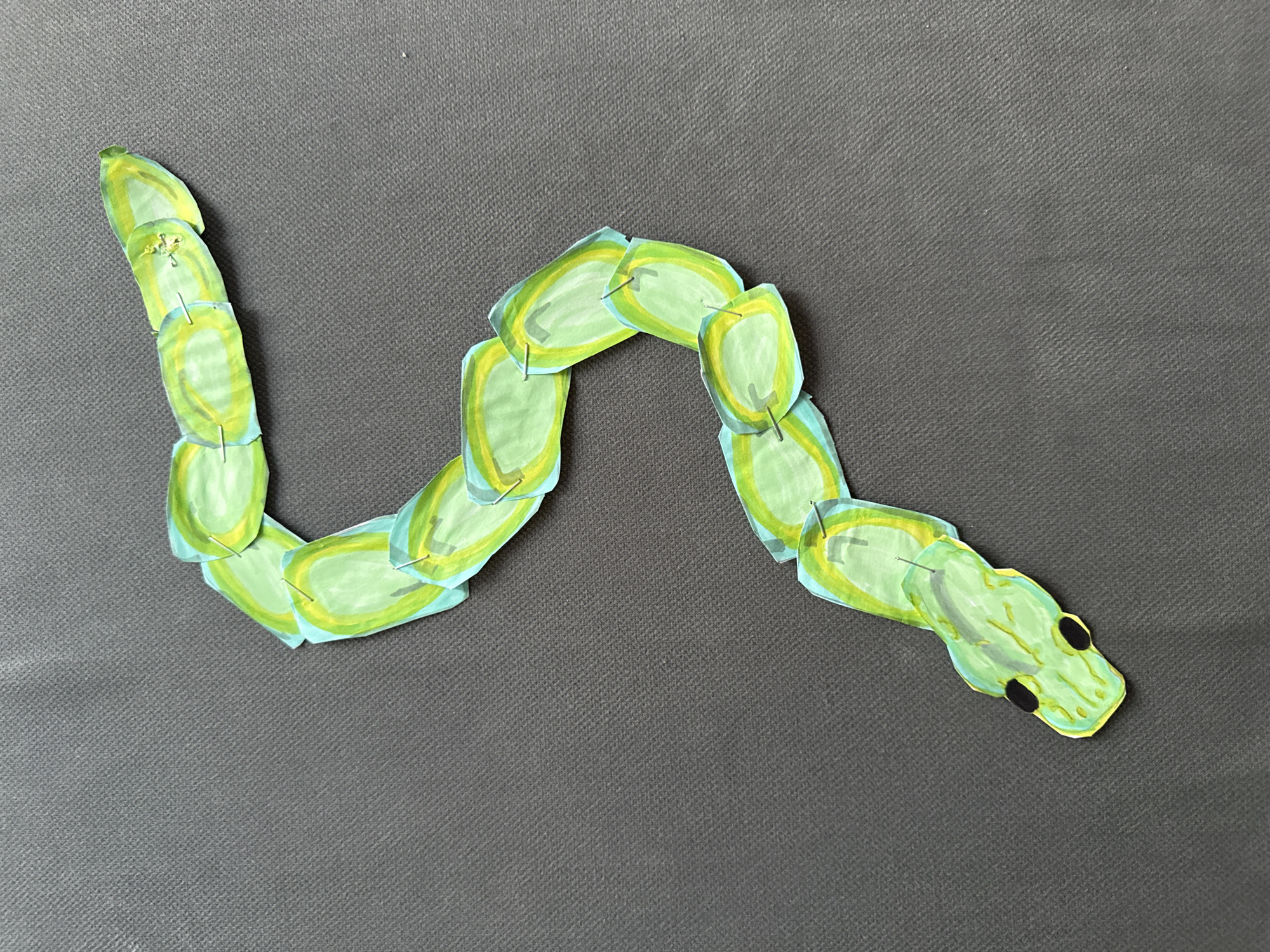 The image displays a handcrafted snake made from paper or cardstock, laid out on a dark gray, textured fabric surface. The snake is composed of multiple segments, each roughly oval or bean-shaped. These segments are predominantly colored in shades of light green and light blue, with some yellow outlines and streaks, giving it a vibrant, stylized appearance.
Each segment is connected to the next by small, thin metal fasteners, likely staples or brads, which are visible at the points where the segments overlap. This construction allows the snake to articulate and form a wavy, serpentine shape across the surface. The snake's head is distinct, featuring a more defined snake-head silhouette with two prominent black circular eyes. The tail end tapers slightly.
The dark gray background has a visible diagonal weave pattern, suggesting a fabric material like felt or a similar textile. There is no specific activity taking place; the snake is a static object artfully arranged. No text is visible in the image.