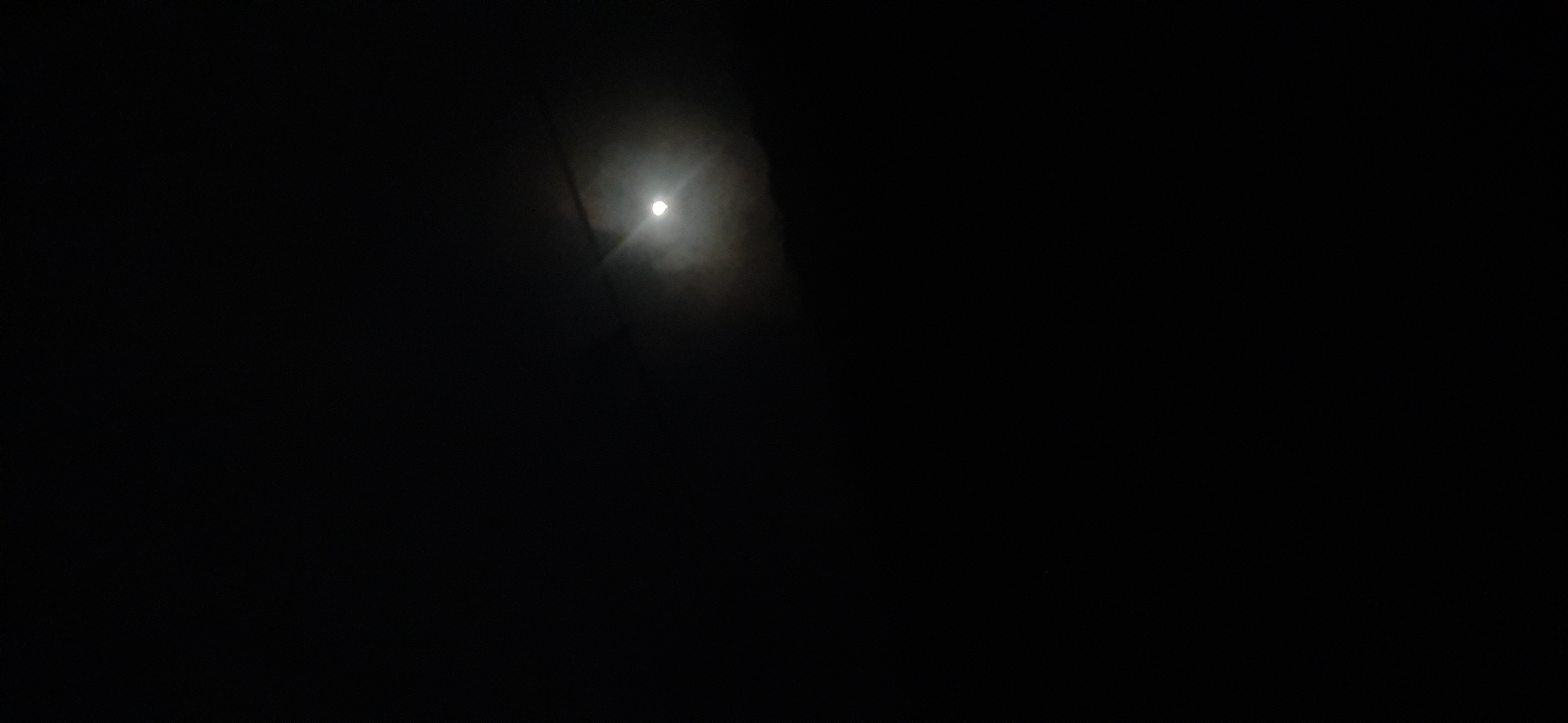 The image shows a bright, full moon in a dark night sky. The moon is slightly obscured by clouds or haze, creating a soft glow around it. There is a subtle lens flare emanating from the moon, indicating a direct light source. The surrounding sky is almost entirely black, with no visible stars or other celestial bodies. The overall scene is stark and atmospheric, suggesting a clear but potentially humid night. There are no discernible structures or natural features of the city of Mūnaişy, Kazakhstan visible in this image.
