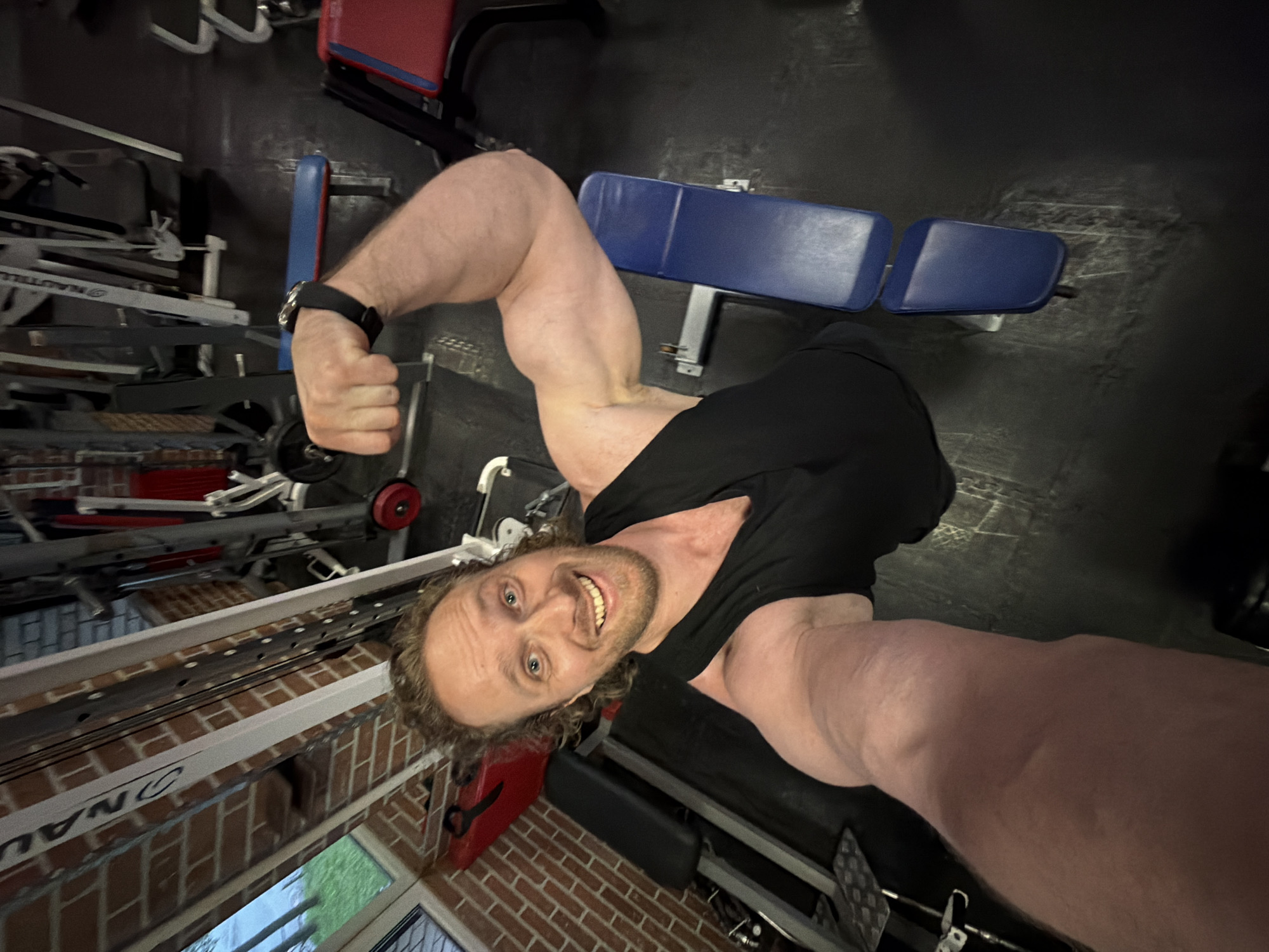 A smiling, light-skinned adult man with curly brown hair and a beard poses for a selfie inside a gym, flexing his prominent right bicep. He wears a black sleeveless top and a black wristwatch on his left wrist. His left arm is extended towards the camera, holding it, with his forearm and hand visible in the foreground. The man’s expression is joyous, with a broad smile directed towards the viewer. Behind him, the gym floor is dark, likely rubber matting. Various pieces of gym equipment are visible, including machines with white and gray frames, pulleys, and cables. A prominent "NAUTILUS" logo is partially visible on a white vertical beam of equipment on the upper left. Multiple upholstered benches in dark blue and red are present. To the left, a reddish-brown brick wall and a window showing green foliage outside are visible. The scene is an indoor gym located in the city of Saint Petersburg, Russia.
