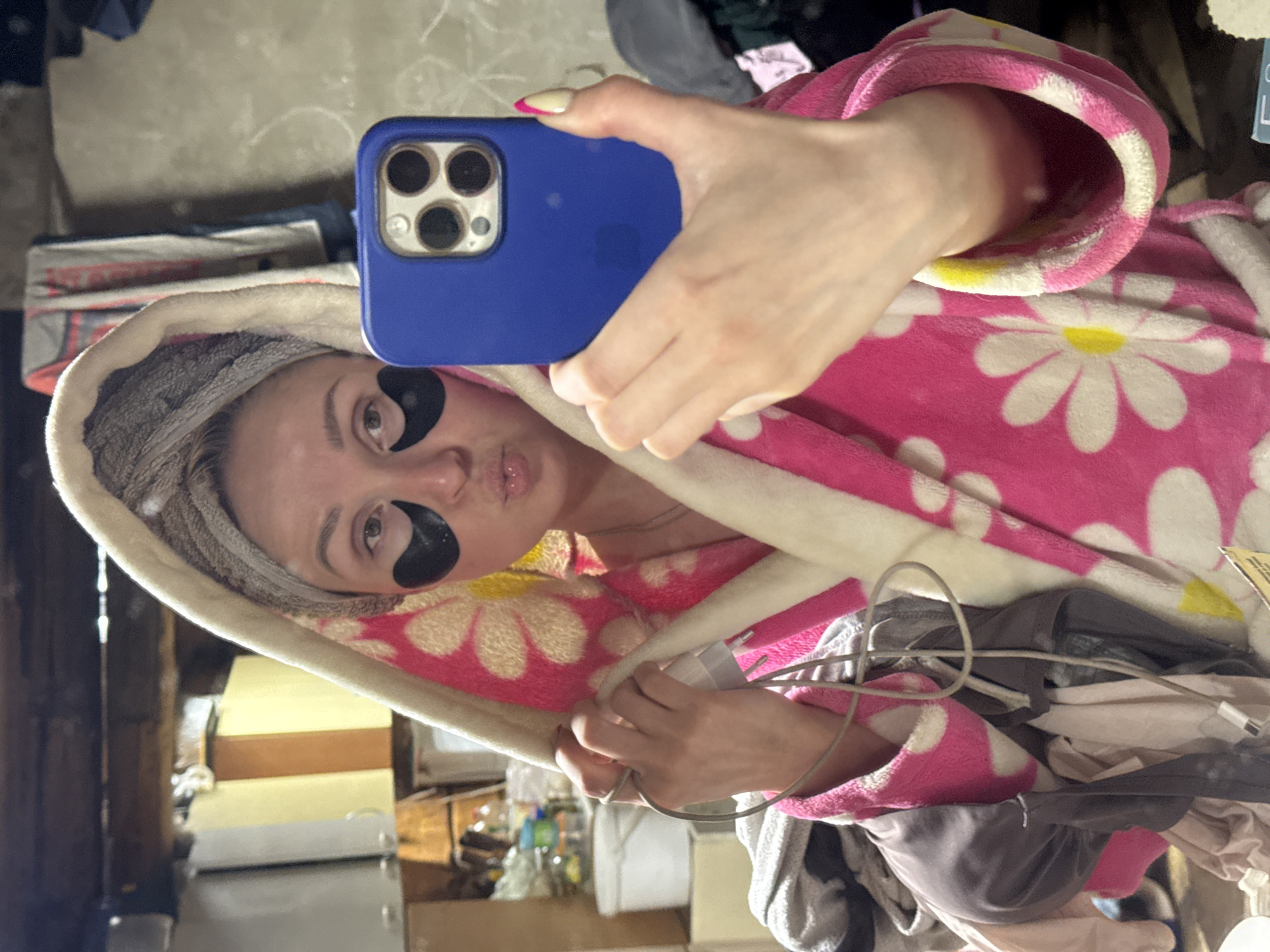 A woman is taking a selfie with a blue smartphone. She has dark patches under her eyes and a towel wrapped around her head. She is wearing a pink robe with white daisies on it. A mess of clothes and wires surrounds her. The background includes a refrigerator and wooden accents. There is no information that indicates this image was taken in Nizhnyaya Chermoda, Russia.