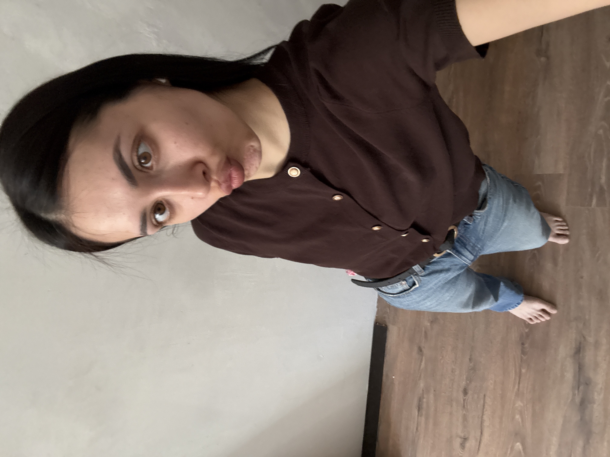A young woman stands indoors, facing the camera. She wears a brown, short-sleeved, button-down shirt and blue jeans with a black belt. Her dark hair is down. She is barefoot. The background consists of a light gray wall and a wooden floor. There is no information in the image to determine the location in the city of Votkinsk, Russia.