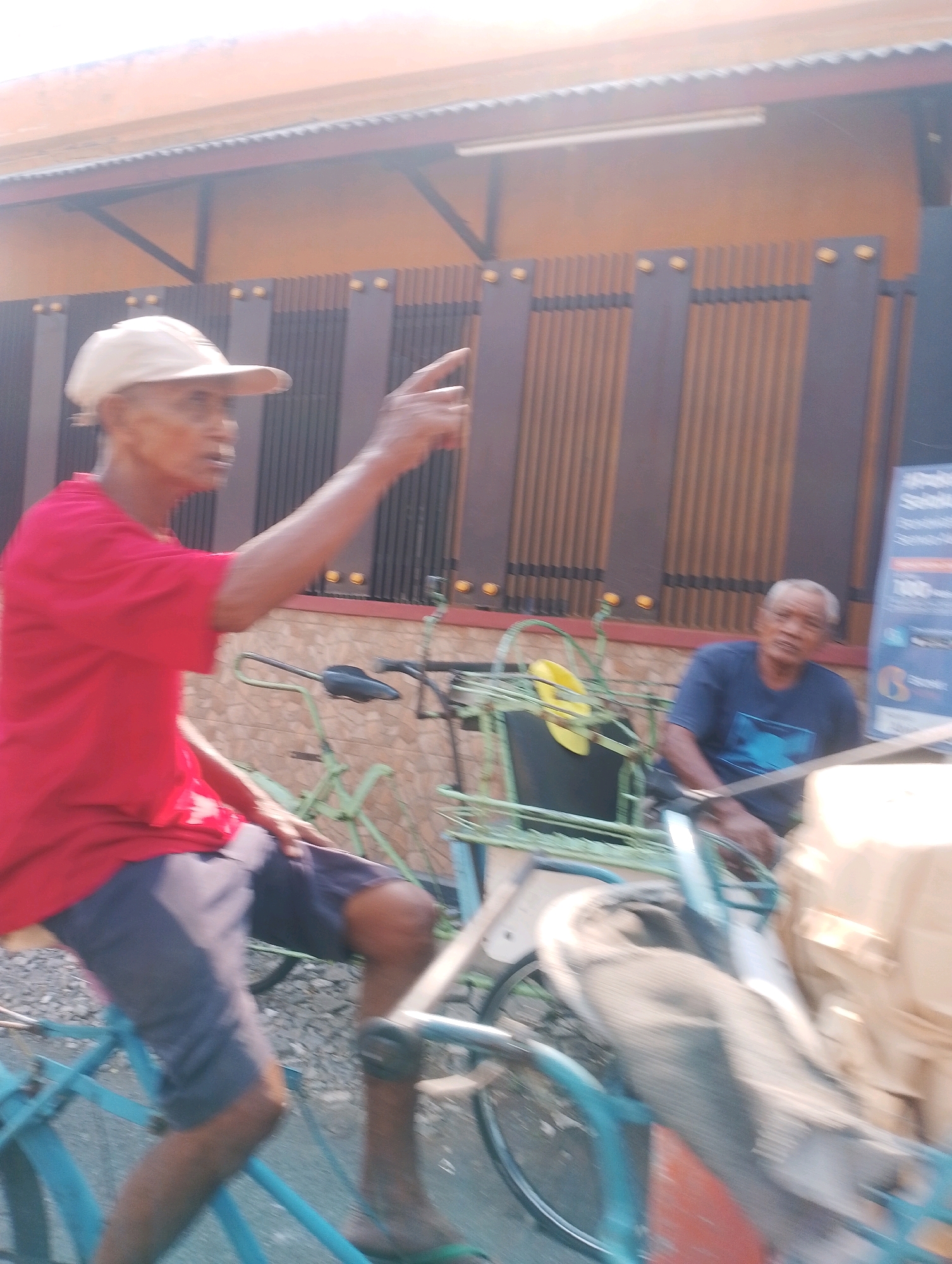 In an outdoor scene on a street in Probolinggo, Indonesia, two elderly men are visible. The man in the foreground, wearing a red t-shirt and a light-colored cap, is riding a turquoise bicycle with a passenger carriage. His left arm is extended, and his index finger is pointing forward, suggesting he is indicating a direction or giving an instruction. He is wearing dark shorts. Behind him and slightly to the right, another elderly man is seated in the passenger carriage of a bicycle that appears to be attached to a larger vehicle. This man is wearing a dark blue t-shirt. The bicycles and carriage are predominantly green and turquoise. In the background, a building with an orange-tiled roof and a dark brown, slatted fence is visible. The lighting suggests it is daytime.