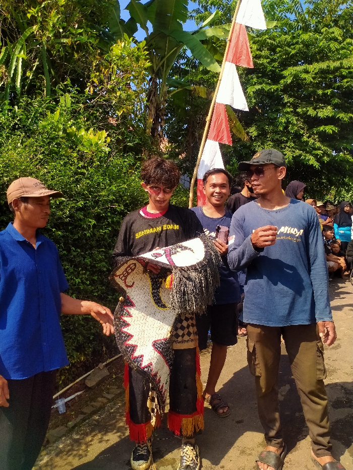 An outdoor scene in Plosorejo, Indonesia, depicts four men standing on an unpaved path under bright, direct sunlight. Lush green foliage, including banana trees, forms a dense background. A bamboo pole on the right supports three triangular flags, alternately red and white.
From left to right:
1. A middle-aged man wearing a brown baseball cap and a short-sleeved blue polo shirt stands with his right hand extended towards the central figure.
2. A young man, likely in his late teens or early twenties, stands centrally, dressed in a black long-sleeved t-shirt with "SENIMAN EDAN" printed in yellow, dark trousers with red and yellow fringes, and dirty white sneakers. His face bears red markings on the cheeks, and he wears dark-rimmed glasses. He holds a large, horse-shaped traditional performance prop, possibly for Kuda Lumping, made of woven material with checkered patterns, red and black accents, and black fringes.
3. Behind the central figure, a young man in a dark blue t-shirt smiles towards the camera, partially obscured by the flag pole.
4. On the far right, a man in a dark baseball cap, dark sunglasses, a long-sleeved blue t-shirt with "MUDA" text, olive green cargo pants, and dark sandals stands with his right hand raised, holding a small dark object. He looks towards the left side of the frame.
A crowd of people is partially visible in the distant background on the right. The ground appears to be a mix of dirt and uneven pavement, with a white PVC pipe section near the lower left.