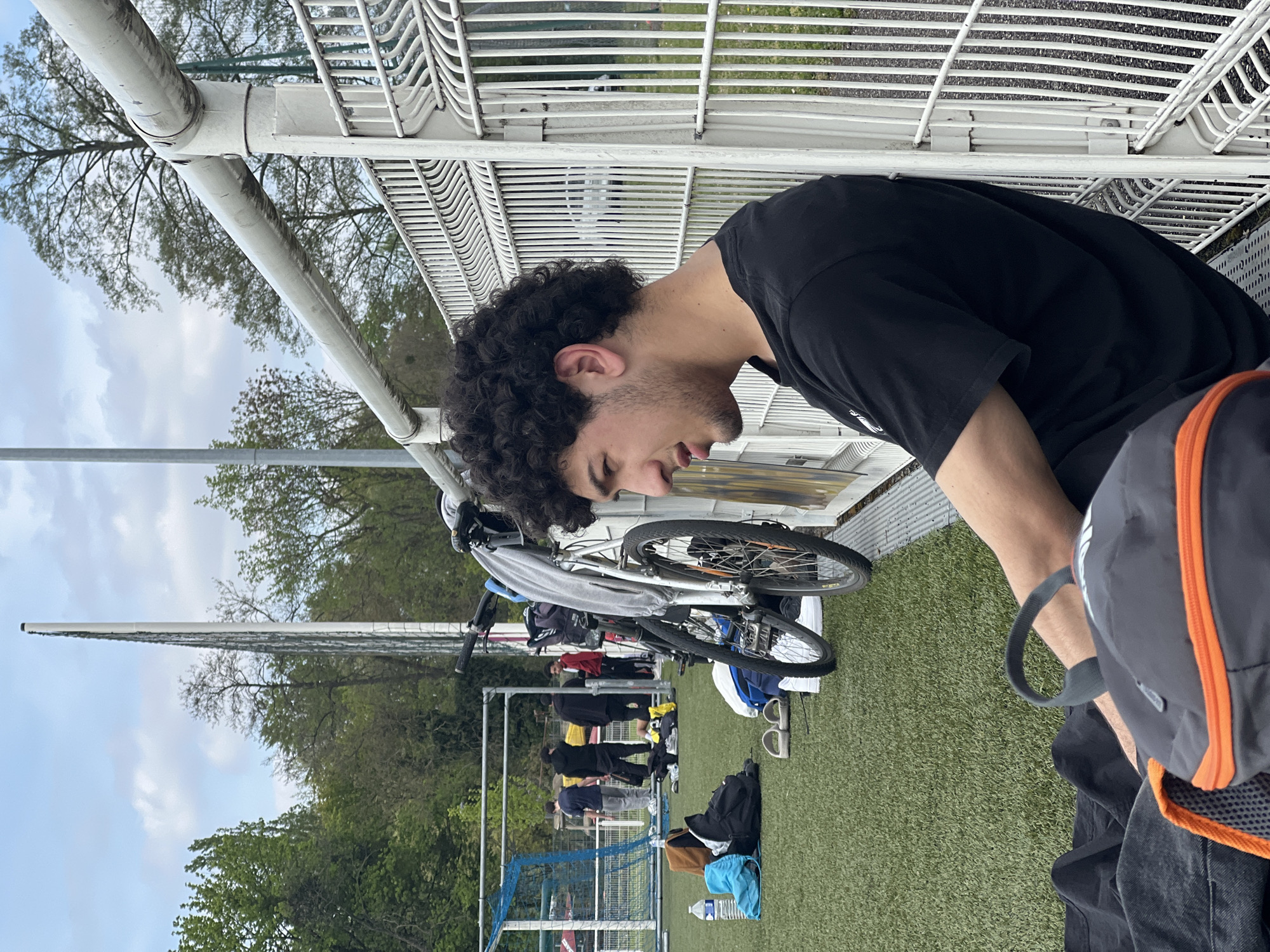 A medium shot, taken from an elevated perspective, depicts a young man with dark curly hair and a black t-shirt, looking down. He is positioned near a white metal structure, possibly a barrier. Behind him, several bicycles lean against the barrier. In the background, a group of people can be seen standing on a grassy field, near what appears to be a soccer goal. The scene takes place outdoors, with trees visible in the distance. There is a bottle of water on the grass. Based on the scene context, the location is likely a sports facility, possibly in the city of Strasbourg, France.