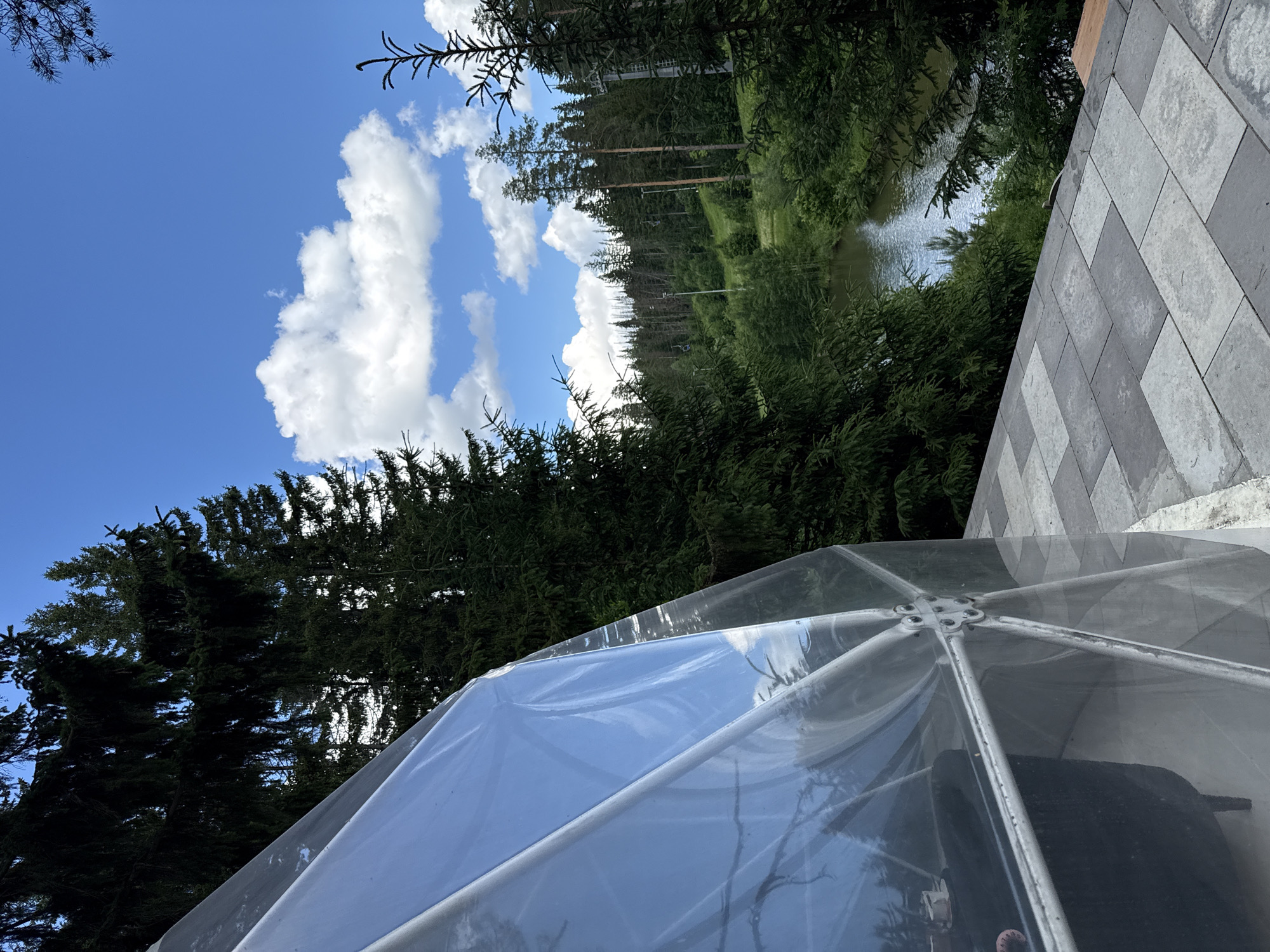 A geodesic dome structure is partially visible on the left of the frame, with a reflective, transparent outer layer. The dome appears to be situated on a paved patio area with large rectangular gray tiles. Beyond the patio, a dense forest of evergreen trees dominates the landscape. A narrow body of water, possibly a river or lake, is visible winding through the trees in the mid-ground. The background shows a hilly terrain covered in a mix of evergreen and deciduous trees, with some dead trees visible. The sky is blue with scattered white cumulus clouds. The scene appears to be located in a natural, possibly mountainous or hilly, area. The location is specified as Nechkino, Russia. There are no people visible in the image.
