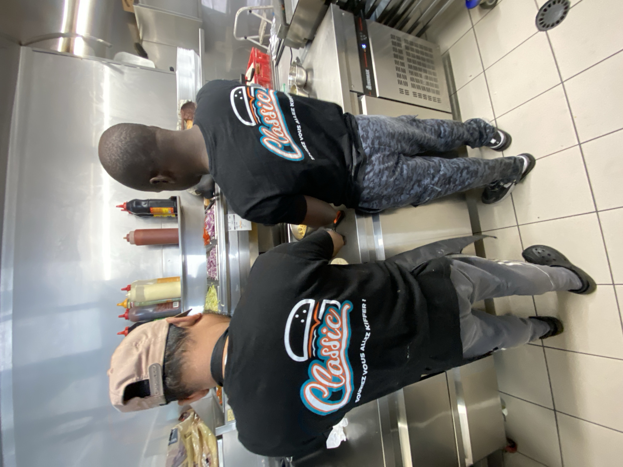 Two men in black shirts with "Classic" written on the back are working in a kitchen. The kitchen is located in Schiltigheim, France. One man is bald and is wearing grey pants with a blue camouflage pattern and black sneakers. The other man is wearing a light pink baseball cap, grey pants, and black Crocs. There are condiment bottles on a shelf in the background. The kitchen has stainless steel surfaces and white tile floors.
