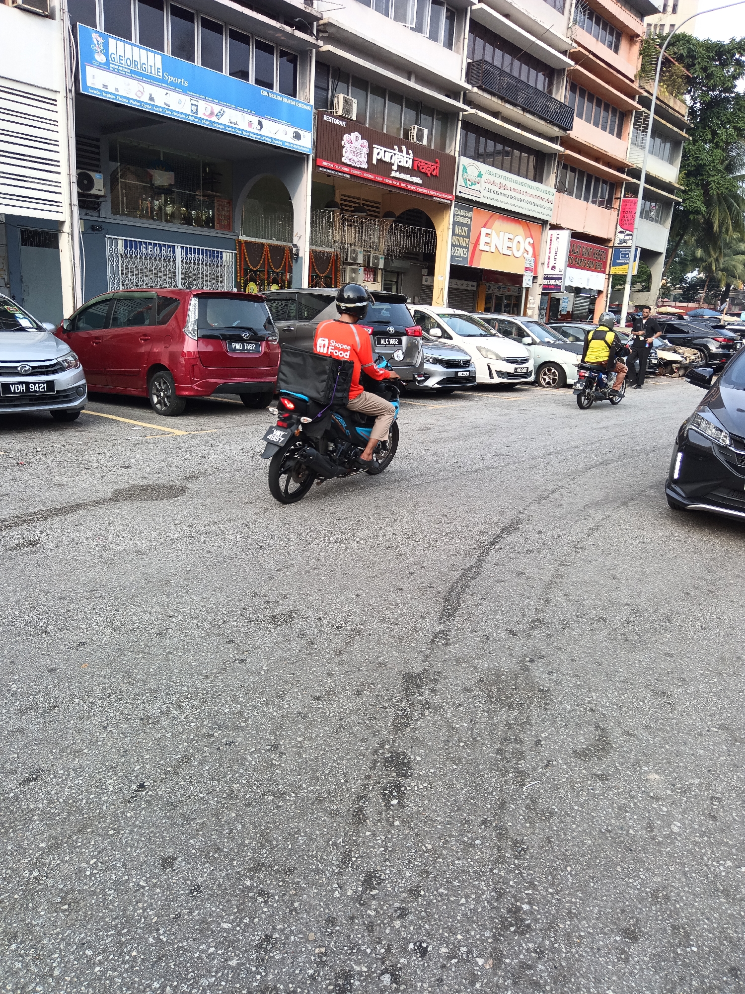 The image captures a street scene in Kuala Lumpur, Malaysia, likely during daylight hours. In the foreground, the asphalt road shows signs of wear and tear with dark stains. A black motorcycle with a rider wearing an orange uniform and a black helmet with a Shopee Food logo on the back is moving away from the viewer. The rider is partially obscured by a large black food delivery box. To the left of the motorcycle, a silver car is parked, and behind it is a red car. Further back, several other cars are parked along the roadside. The background features a row of multi-story buildings with shops on the ground floor. Signs on the buildings indicate various businesses, including "GEORGE TE Sports," "Punjabi Rasoi," "PERSATUAN PENGURUSAN KESELAMATAN INDUSTRI MALAYSIA," and "ENEOS." A street lamp is also visible. The environment appears to be a commercial area with moderate vehicular traffic.