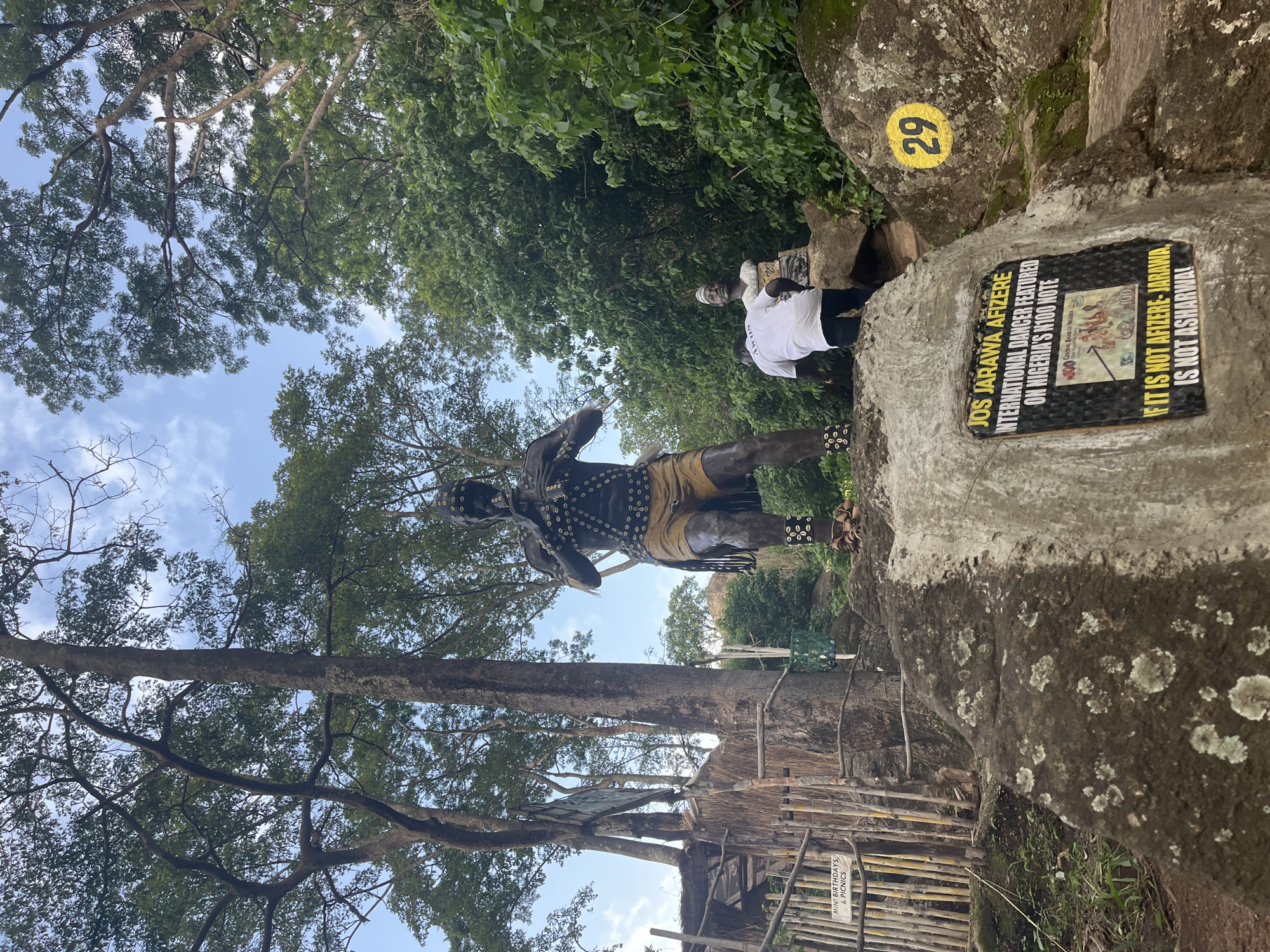 The media file shows a statue of a dancer, likely in an outdoor park or historical site. The statue is adorned with beads and a headdress, and is positioned in front of a large rock formation. To the right of the statue, there is a sign that reads "JOS JARAWA AFIZERE INTERNATIONAL DANCER FEATURED ON NIGERIA'S N100 NOTE IF IT IS NOT AFIZERE-JARAWA IS NOT ASHARWA!". There is also a yellow circle with the number "29" painted on the rock. In the background, there are trees and lush greenery, suggesting a tropical or subtropical environment. A person in a white shirt and black pants is visible near the rocks, possibly a visitor or guide. The sky is visible through the trees, indicating it is daytime. The scene appears to be a cultural or tourist attraction.