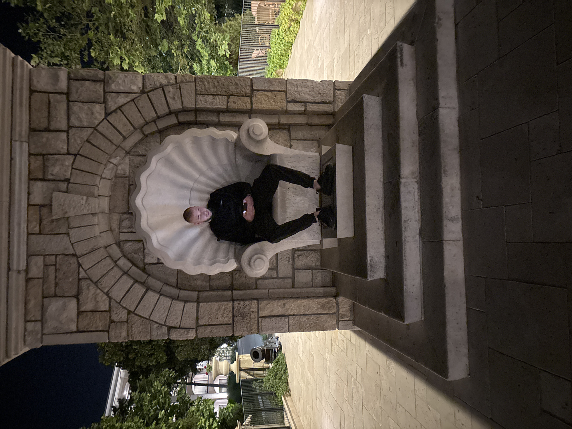 A young man in a black hooded sweatshirt and black pants is seated on a large, seashell-shaped stone throne. He is looking down at his phone, which he holds in his hands. The throne is situated within a stone alcove that features an arched opening and is adorned with rough-hewn stone blocks. In front of the throne are three wide stone steps leading down to a paved walkway. The scene is illuminated by artificial lighting at night. To the left of the throne, a building with white facade and green trees are visible, along with a black metal fence and a decorative urn. To the right, there is more greenery and a metal fence. The location is the Sea Garden in Varna, Bulgaria, near the Alley of First Love.
