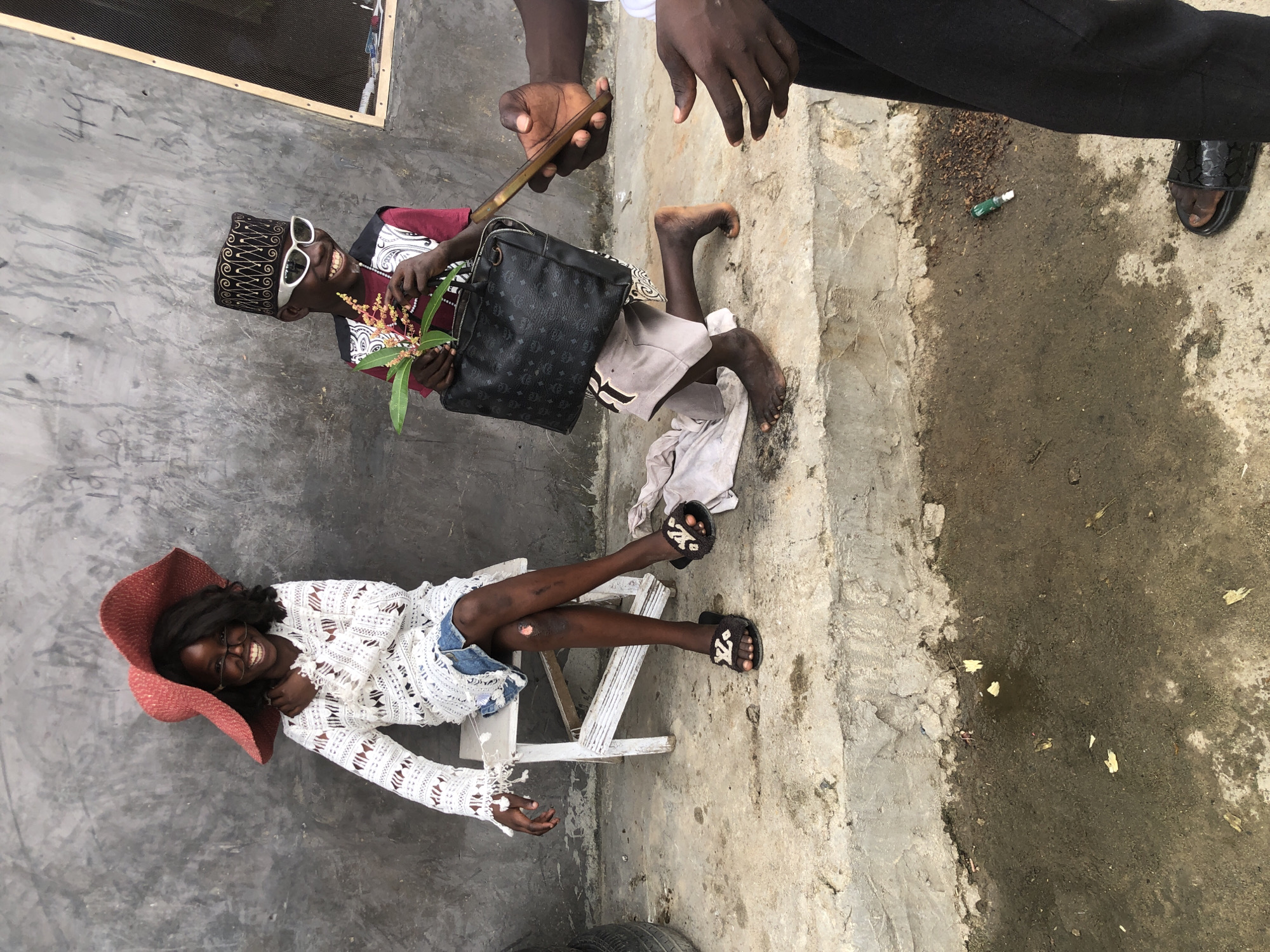 Two children are posing for a photo in what appears to be an outdoor urban setting in Ado, Nigeria. The child on the left is seated on a white stool, wearing a large red hat, glasses, a white lacy dress over denim shorts, and embellished slide sandals. The child on the right is kneeling, wearing a decorative cap, white sunglasses, a vest, and a red shirt, holding a branch with flowers and a black handbag. Another individual's hands are visible, one holding a smartphone, suggesting a photograph is being taken. The background is a rough, textured concrete wall. The ground is concrete with some puddles of water and debris.