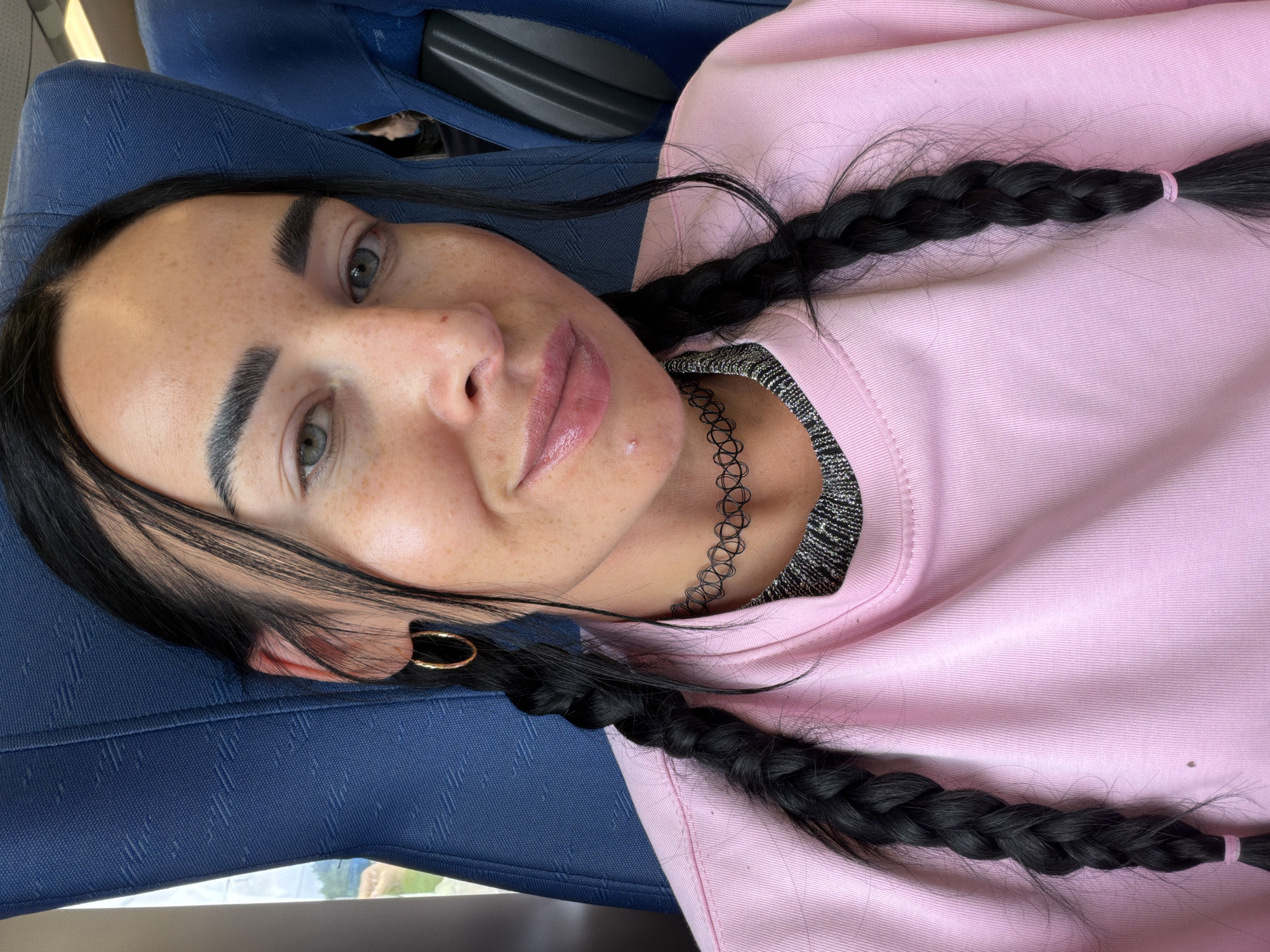 A close-up, vertical shot features a person with fair skin and dark hair styled in two braids. Their eyes are open, and they are looking towards the camera. The person has freckles across their nose and cheeks. They are wearing a light pink sweatshirt and a black choker necklace. A gold hoop earring is visible in their left ear. The background is a textured blue surface, possibly a seat or upholstery, suggesting an indoor setting like a vehicle. The lighting indicates it is daytime, with natural light entering from the left side of the frame. The image conveys a casual and relaxed mood. There is no visible text in the image.