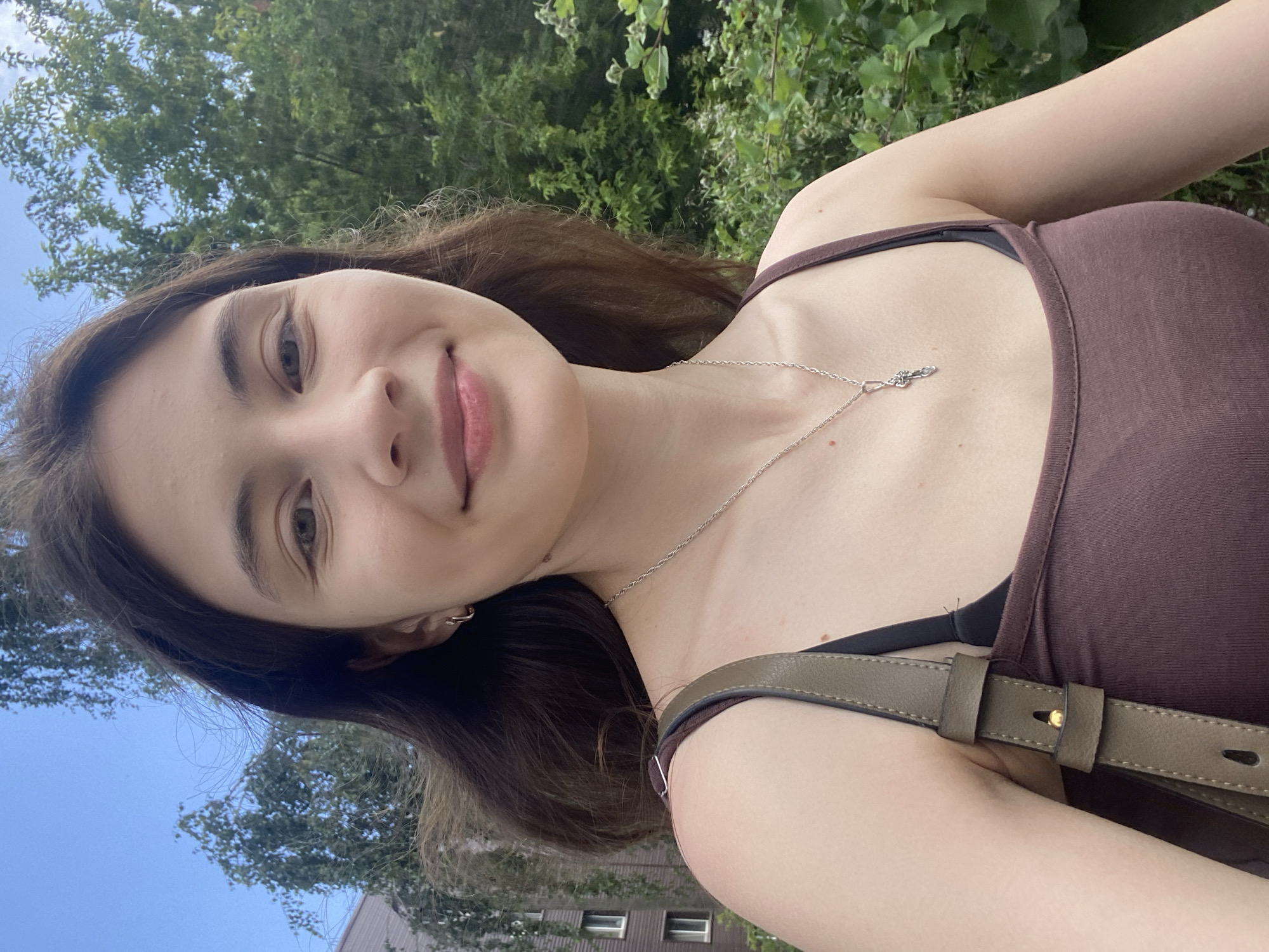 An outdoor selfie shows a young woman with long brown hair and light skin. She is wearing a brown tank top and a silver necklace with a key pendant. Her right ear is adorned with a small hoop earring. A portion of a green handbag strap is visible on her left shoulder. Behind her, a blurry background reveals lush green foliage and trees, with a hint of a building on the far left. The sky above is a clear blue. The scene appears to be set in a park or residential area, possibly in Votkinsk, Russia, given the context.