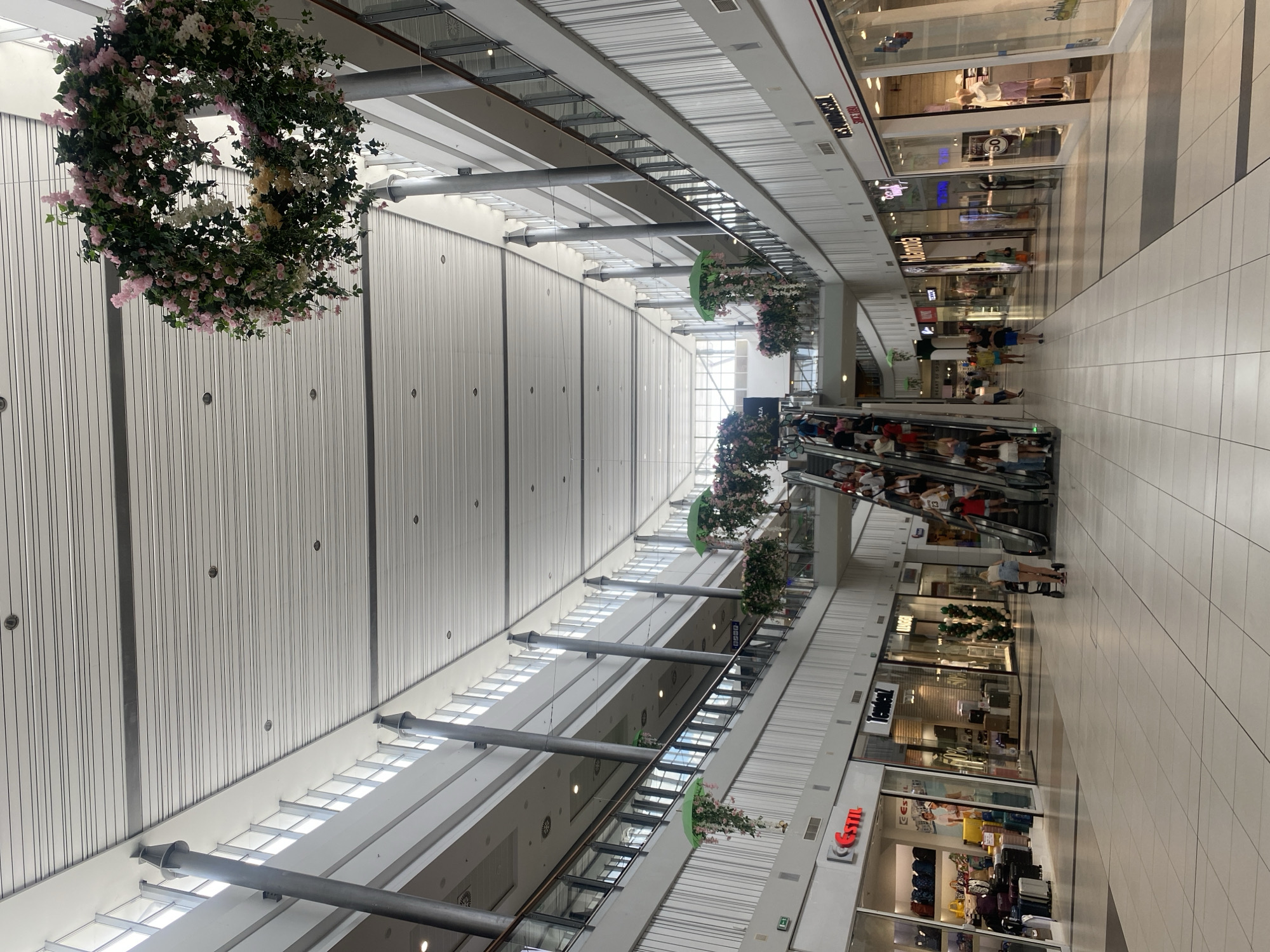 This is a wide shot of the interior of a multi-level shopping mall, likely the Galleria Burgas in Burgas, Bulgaria. The architecture features a high, sloped ceiling with a metallic, ribbed surface, crisscrossed by structural beams and lit by recessed ceiling lights and natural light filtering through large windows along the sides. Several cylindrical support columns, painted a dark gray, run vertically throughout the space. Suspended from the ceiling are large, circular floral arrangements, one prominent in the upper center of the frame, and several smaller, umbrella-shaped floral decorations hanging along the upper-level walkways.
The mall's ground floor and upper levels are visible, with shops lining both sides of the wide, open central walkway. The floor is tiled in a light-colored, checkered pattern. A busy escalator is situated in the middle of the mall, with numerous people ascending and descending. Shoppers, dressed in casual summer attire, are spread throughout the scene, some walking, others on the escalator, and a few are visible in the shop windows displaying clothing and merchandise.
On the ground floor, several shop fronts are identifiable. To the left, a store labeled "GSTIL" is visible, with displays of luggage and handbags. Further down, other shops with illuminated signs are present, including one with a large "50%" sign in its window. The upper levels feature glass balustrades along the walkways, offering views of the lower levels. The overall impression is of a modern, well-lit, and active retail environment.