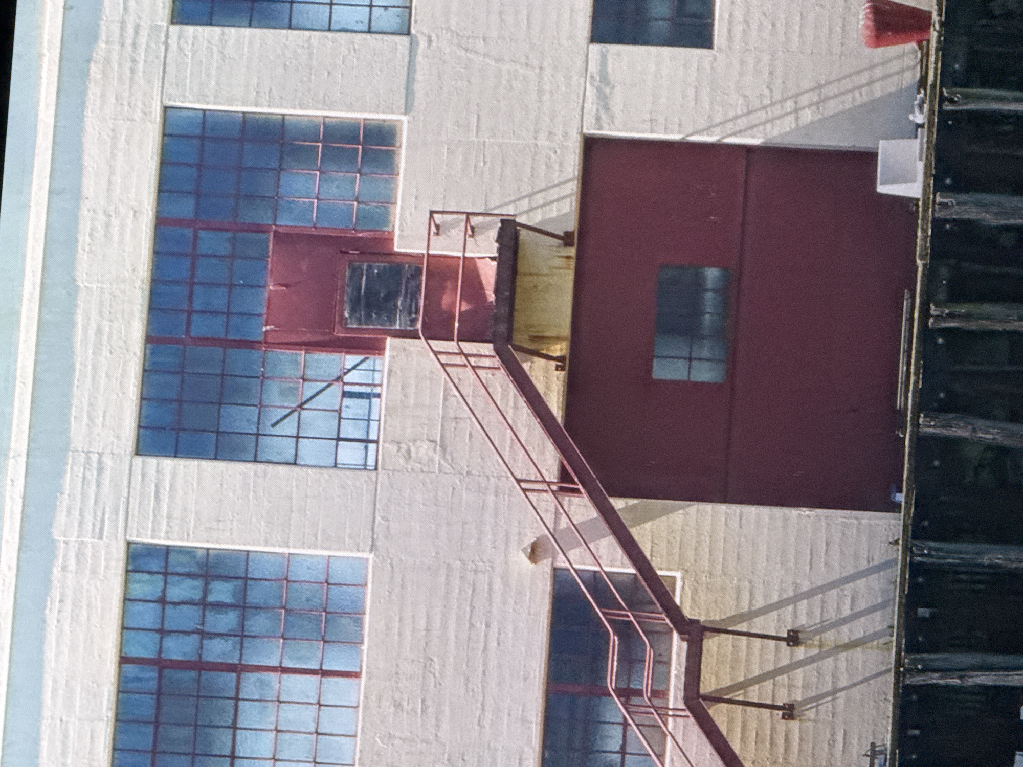 A weathered, light-colored brick building with large, multi-paned windows is visible. A red metal staircase with a railing ascends the side of the building, leading to a red door with a dark window panel. The building has sections of dark red siding. The scene appears to be near a body of water, with wooden pilings visible in the foreground. The image was taken on a bright, sunny day, casting distinct shadows. No people are present. The architectural style and condition suggest an industrial or commercial building, possibly a warehouse or factory. The location is identified as being in Saratov, Russia.