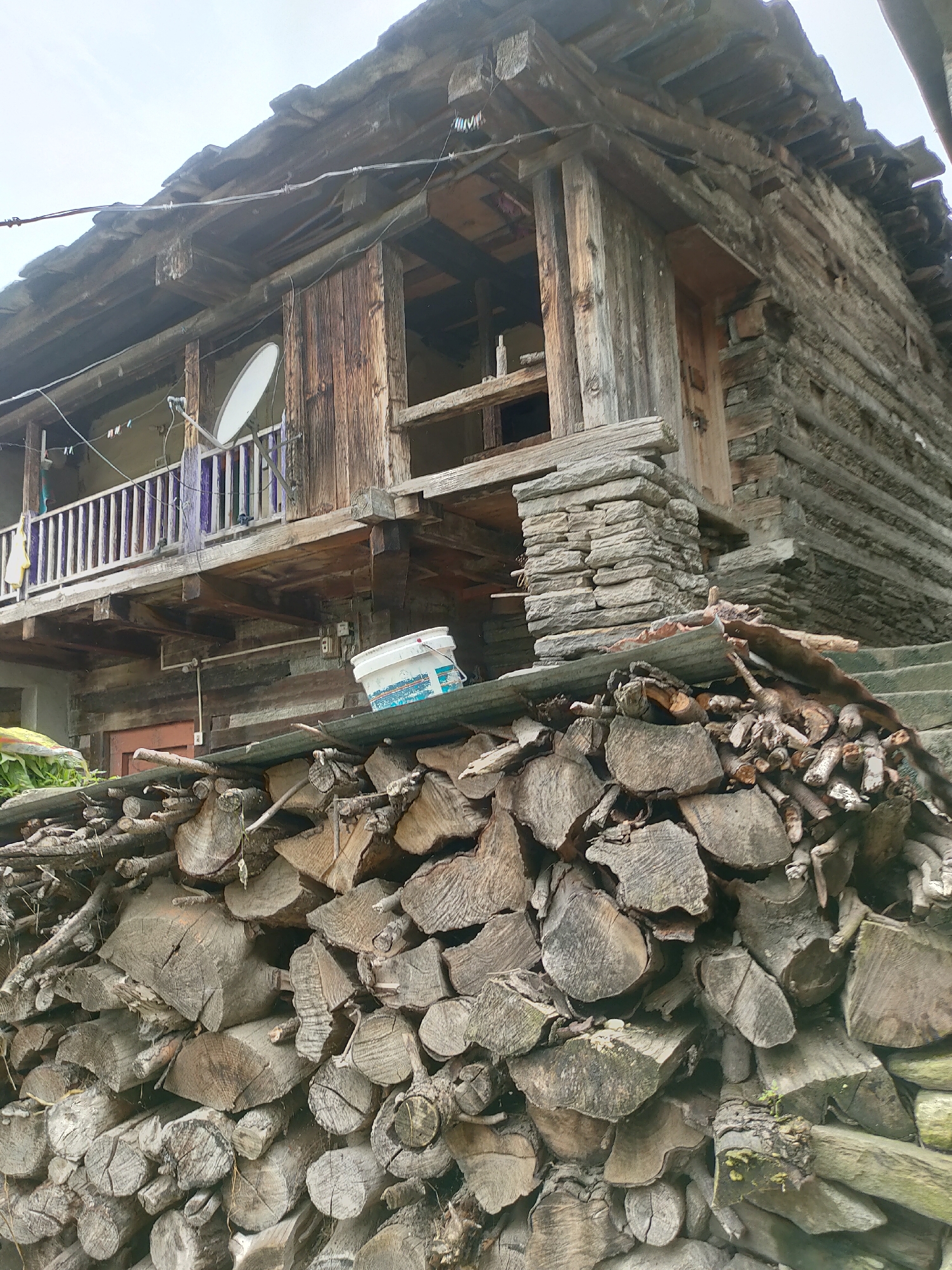 This image depicts a rustic, wooden structure, likely a house, in Damadi, India. The building is constructed from rough-hewn logs and features multiple levels, balconies with railings, and a sloped, wooden roof covered with what appear to be stone slabs. A white satellite dish is mounted on the exterior of the upper level. In the foreground, a large stack of chopped firewood is piled high, with various sizes and types of logs visible. A white bucket sits on a slanted surface above the firewood, near the base of the building. The overall scene suggests a rural or mountainous setting, with the architecture and the presence of firewood indicating a traditional way of life. The sky is overcast. There are no people visible in the image.