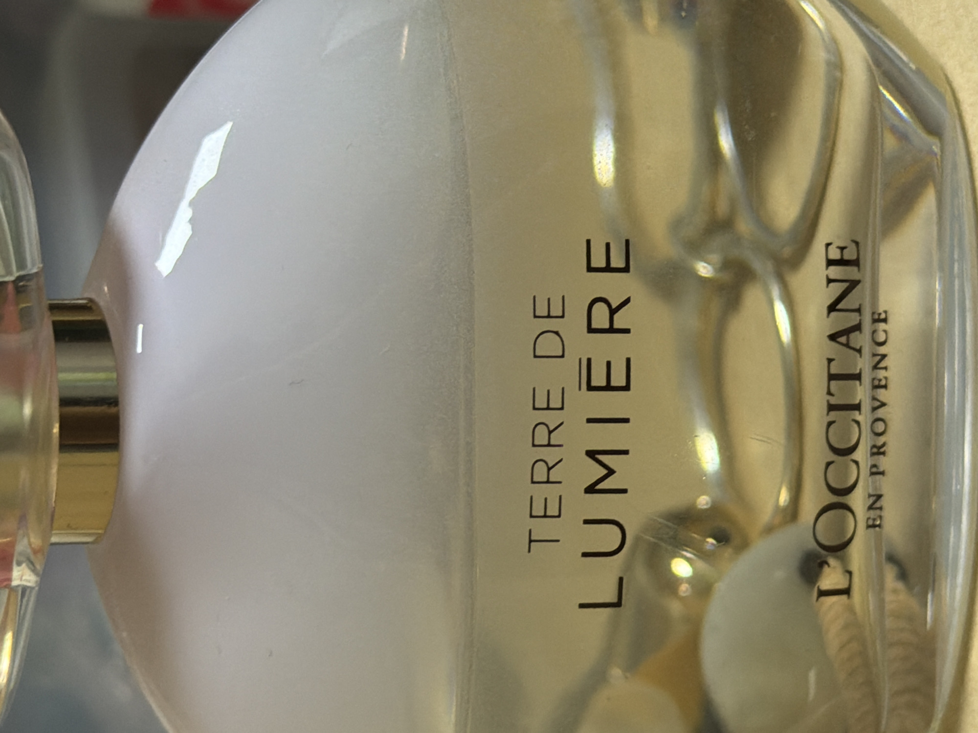 The image shows a close-up of a bottle of L'Occitane Terre de Lumière perfume. The bottle is made of glass and contains a light yellow liquid. The brand name "L'OCCITANE" and the product name "TERRE DE LUMIÈRE" are printed on the bottle in black lettering. The background is out of focus, but appears to be an indoor setting, possibly a store or a vanity. There is no indication of the location being Bishkek City, Kyrgyzstan.