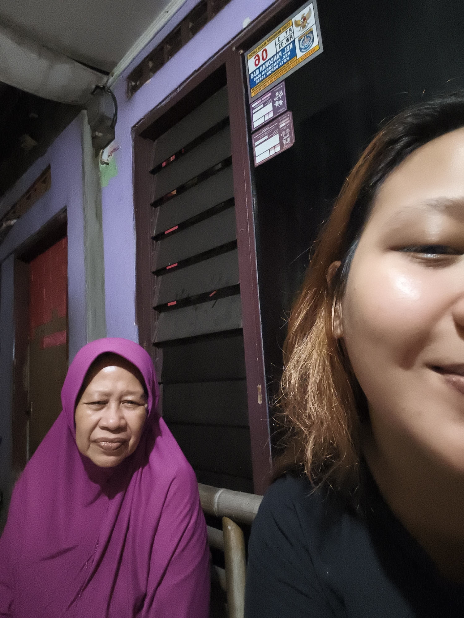 Two individuals are in the frame: an elderly woman on the left wearing a magenta hijab and a younger individual on the right, partially visible, in a black shirt. They are likely outside a building at night. A sign with Indonesian text and emblems is attached to the building above and behind them. The building has a lavender exterior with a louvered window or door. There is a door visible to the left, painted red inside and lavender on its exterior frame. The environment is dimly lit. The sign appears to read "30" and refers to "KEL. PANCORAN MAS" and "KEC. PANCORAN MAS," indicating a location in the Pancoran Mas district of Depok, Indonesia.