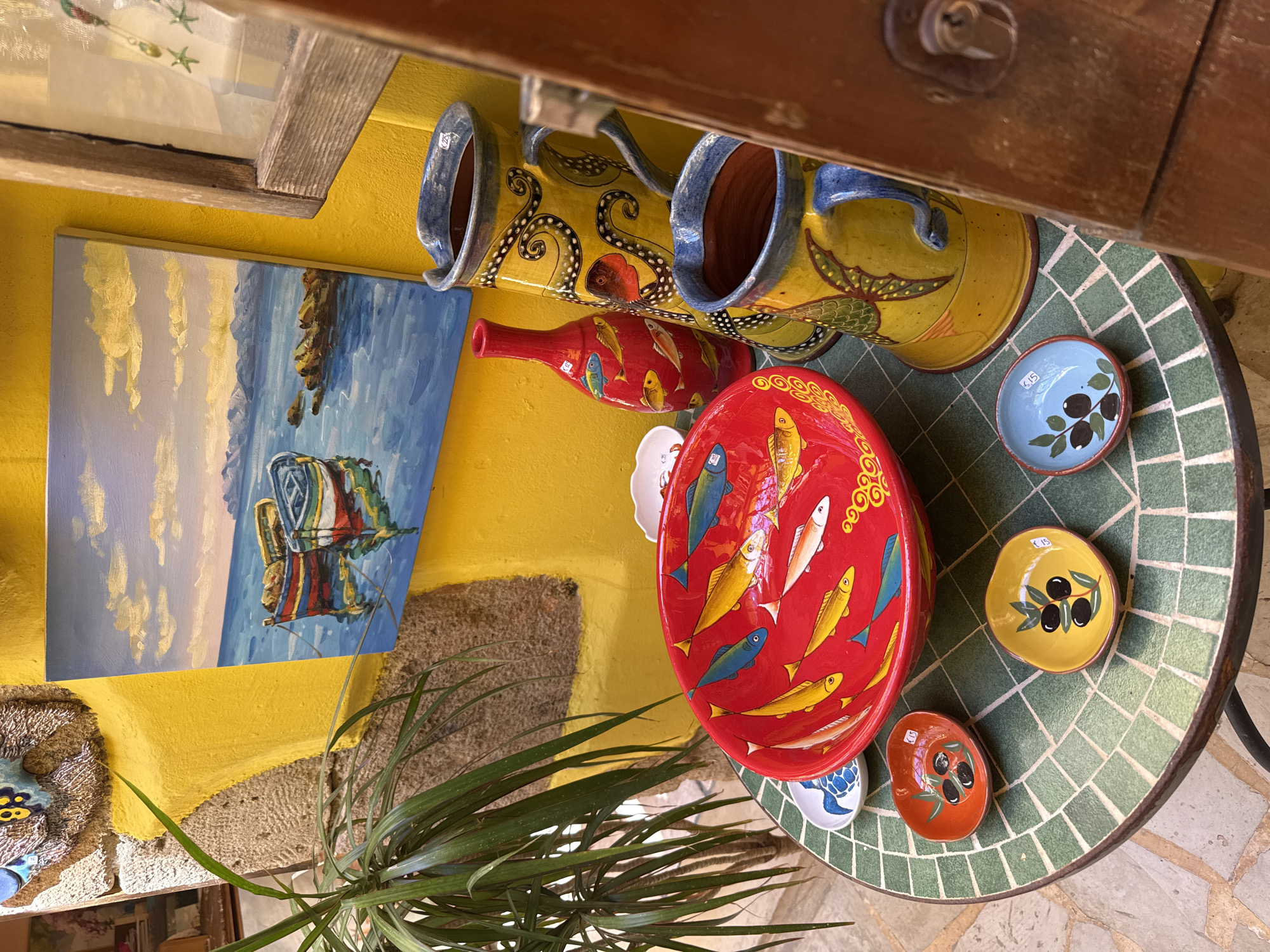 A still life display of decorative ceramic pottery and a small painting is arranged on a mosaic-tiled table outdoors, likely in the city of Chania, Greece, given the style of the pottery and architecture. The scene is bathed in sunlight.
In the foreground, a round, bright red ceramic bowl is prominently featured, filled with various painted fish in shades of yellow, blue, and white. To the left of this large bowl, a small white dish with a blue turtle design is partially visible. Arranged around the larger bowl are three smaller, oval-shaped dishes. The dish closest to the viewer is orange with black olives and green leaves. To its right is a yellow dish with a similar olive branch design. The third small dish, to the right of the yellow one, is light blue with black olives and green leaves.
Behind the table, a tall, slender red ceramic vase with painted fish is placed. To the right of the vase stand two large, yellow ceramic pitchers decorated with octopus tentacles and other marine-themed imagery. The pitcher in the foreground has a blue rim and handle, while the one behind it also has a blue rim.
Above the table and pottery, a vibrant yellow wall serves as the backdrop. Mounted on this wall is a small, rectangular painting depicting three colorful boats floating on a blue sea under a sky with fluffy clouds and distant mountains. To the far right, a wooden door or frame is visible, with a part of a window showing some decorative items. To the left of the yellow wall and painting, a section of rough-hewn stone wall and a green leafy plant add texture to the scene. The table itself has a surface composed of small, square green mosaic tiles, with a darker metal base. The ground surrounding the table is paved with irregular stones.