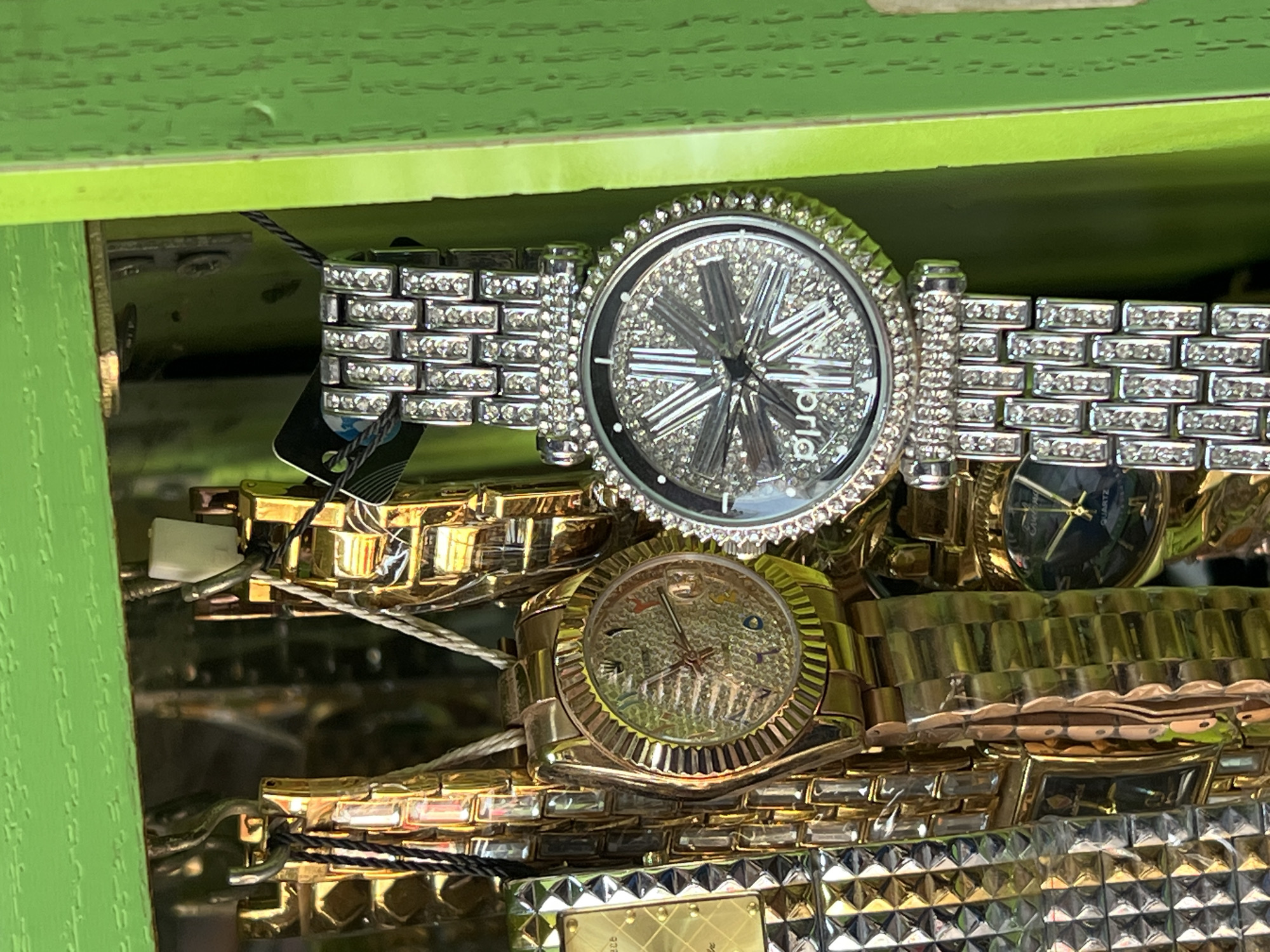 In Bukuru, Nigeria, a close-up, eye-level shot captures a collection of watches displayed within a bright green, compartmentalized shelf unit. The watches are suspended from hooks, with some featuring gold-toned bands and dials with Arabic numerals, while others boast silver-toned bands and faces encrusted with rhinestones. One prominent gold watch on the left has a textured gold dial. To its right, a silver watch features a dial with a starburst pattern of rhinestones. The watches are neatly arranged, suggesting a retail or display setting. The background is slightly blurred, but the green shelving unit is distinct.