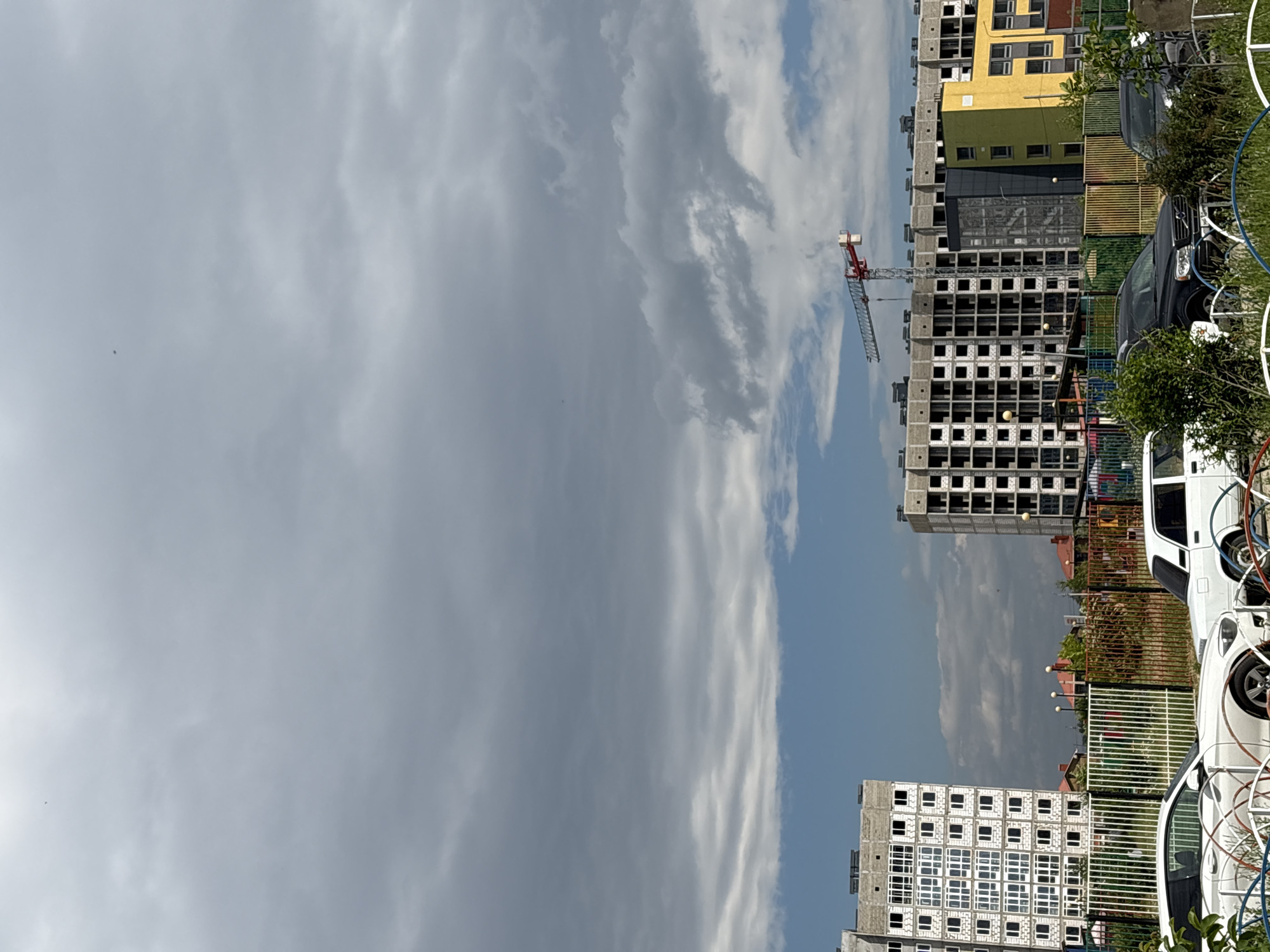 This image depicts an overcast sky above a construction site in Mikhaylovsk, Russia. In the foreground, two white cars are parked, partially obscured by a fence. To the left, an unfinished multi-story building with many windows is visible. In the center and right, another multi-story building, also under construction, dominates the scene, with a construction crane positioned above it. The buildings are modern in design, with visible concrete structures and blank window openings. The environment appears to be a developing urban area, with other residential buildings and greenery visible in the background. The overall atmosphere is one of ongoing development and construction under a gray, cloudy sky.