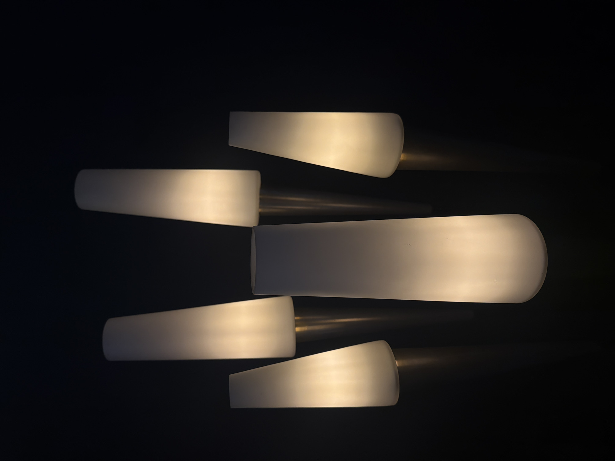 The image shows an indoor light fixture. The fixture contains five frosted white glass shades, each attached to a cylindrical metal base. The shades vary in length. The background is black. The location is not apparent from the image.