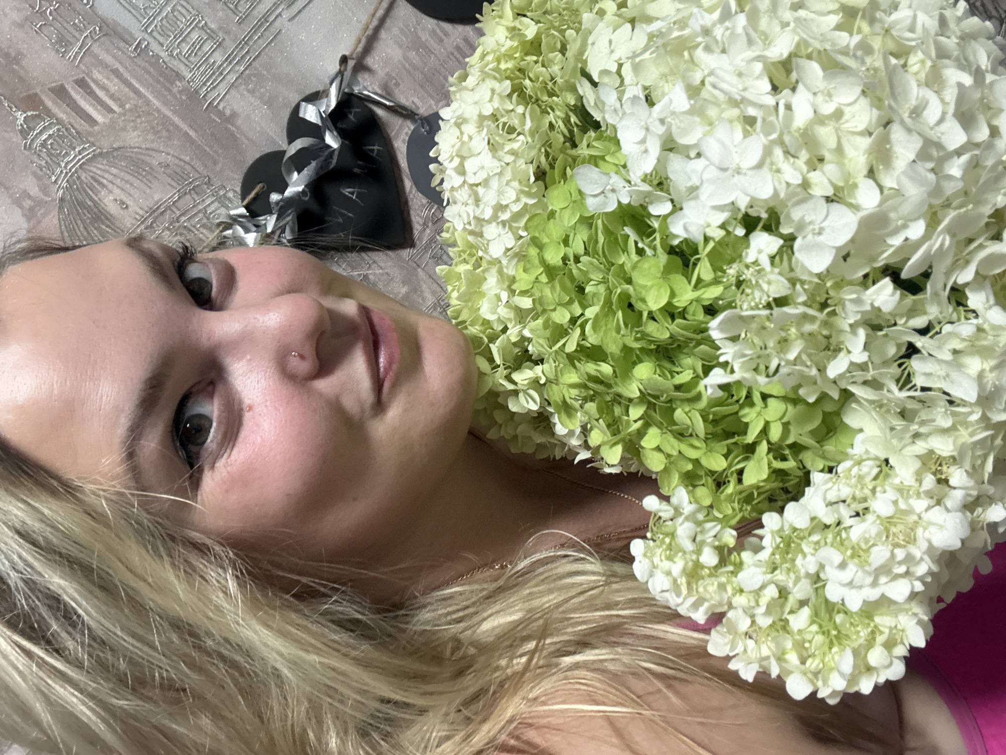 In the foreground, a woman with blonde hair and a pink top is holding a large bouquet of white and green hydrangeas. She is looking at the camera with a slight smile. The background shows a patterned wall with what appears to be a decorative element of several black heart shapes with silver ribbon. The woman has a small nose ring and a delicate necklace. The scene is indoors.