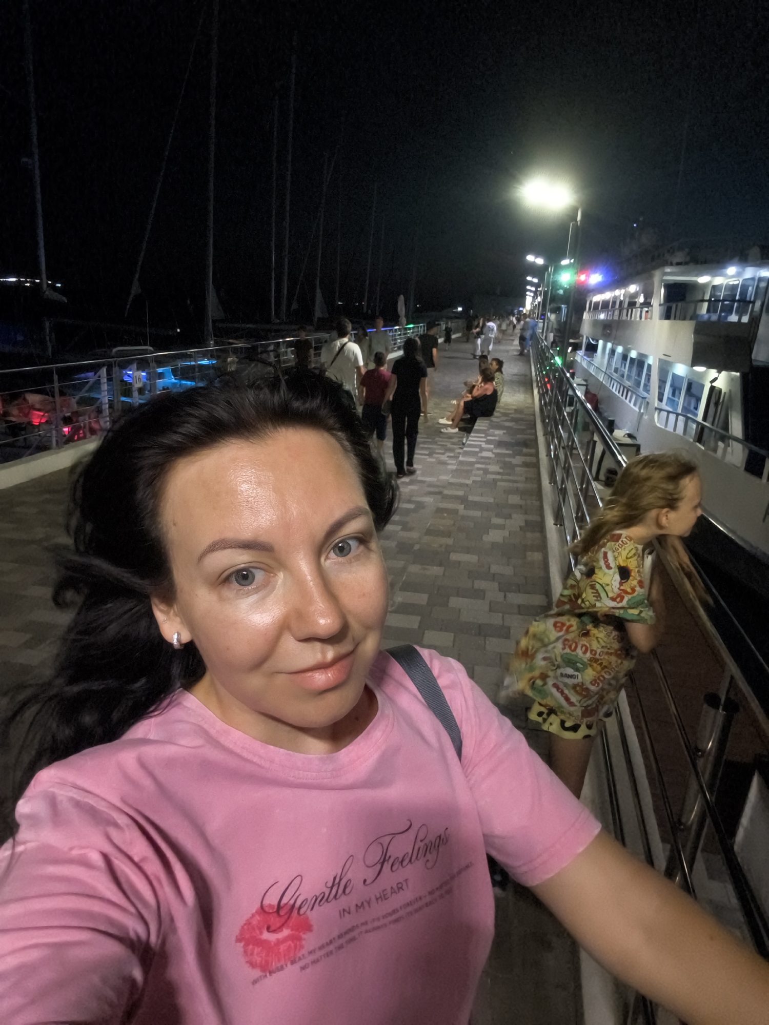 A woman in a pink t-shirt takes a selfie on a paved promenade at night. She has dark hair and light eyes. In the background, there are several people walking, a large white boat docked, and masts of sailboats. The promenade is illuminated by streetlights, and there are railings along the edge overlooking the water and boats. The woman's t-shirt has a graphic of a kiss and text that reads "Gentle Feelings IN MY HEART". Another woman, in a colorful patterned dress, is leaning on the railing in the background, looking at the boat. The location appears to be a harbor or marina.