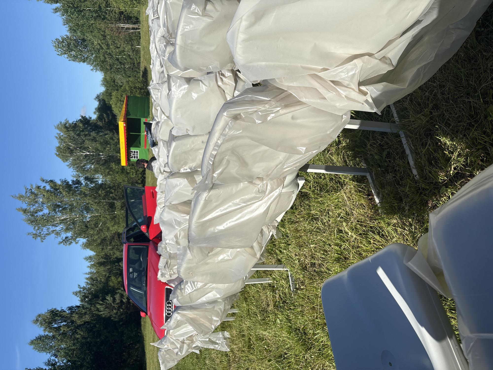 The image shows an outdoor scene on a sunny day with a clear blue sky. The foreground is dominated by a large pile of white, translucent material, likely plastic sheeting, draped over tables. The sheeting is crinkled and folded, suggesting it's being stored or prepared for an event.
To the left of the sheeting, a red car is visible with its doors open. The car appears to be parked on a grassy area. Further in the background, a small, colorful structure, resembling a vendor booth or a small amusement ride, is visible. It has a green body with a yellow and red striped roof. Behind the booth, a line of trees forms a dense backdrop against the sky.
A person is partially visible near the colorful structure, wearing a dark shirt. The overall impression is of a setup for an outdoor event, possibly a fair or market, taking place in a rural or semi-rural setting in Belarus, as indicated by the location context. The time of day is clearly daytime due to the bright sunlight casting shadows, and the weather appears to be fair and warm. There is no visible text in the image.