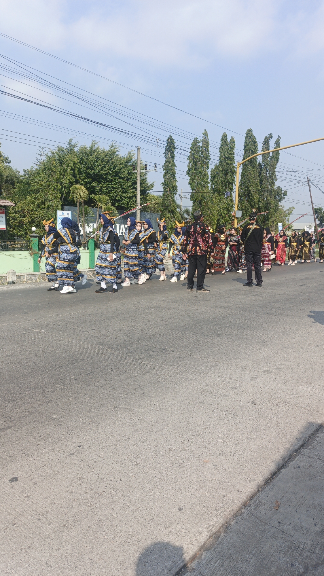 In Sumengko, Indonesia, a group of individuals in traditional attire are participating in a parade. The parade is taking place on an asphalt road with a slight incline. Several trees line the background, and power lines are visible overhead. The participants, dressed in matching blue and white patterned outfits with headwear, are walking in a procession. Some are carrying what appear to be decorative poles. A few individuals in the foreground are wearing different, more ornate outfits. The sky is clear and blue, indicating a sunny day. The scene suggests a cultural event or celebration in the town.