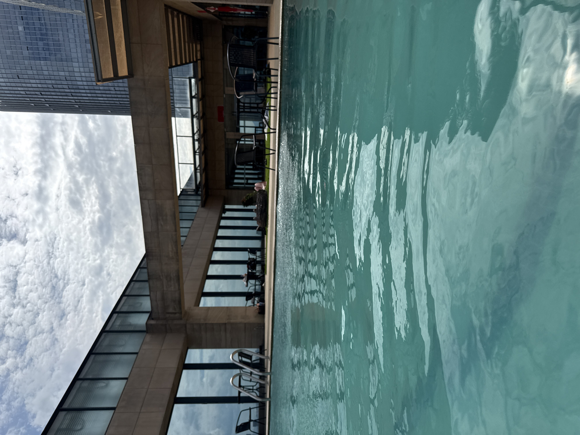The image shows an outdoor swimming pool on a cloudy day, likely located on a rooftop or elevated level of a building in Yiwu, China. The foreground is dominated by the turquoise water of the pool, with ripples and reflections indicating movement. In the background, a modern architectural structure with large glass windows and concrete elements frames the scene. Beyond this structure, a tall, dark glass skyscraper, characteristic of a city skyline, is visible. Several empty black chairs and tables are placed on a tiled area adjacent to the pool, suggesting an outdoor seating area. A pair of silver pool ladders are positioned at the edge of the water. In the distance, a few figures are seated at tables, and one person appears to be lounging near a rock formation. The sky is filled with a mix of white and gray clouds.