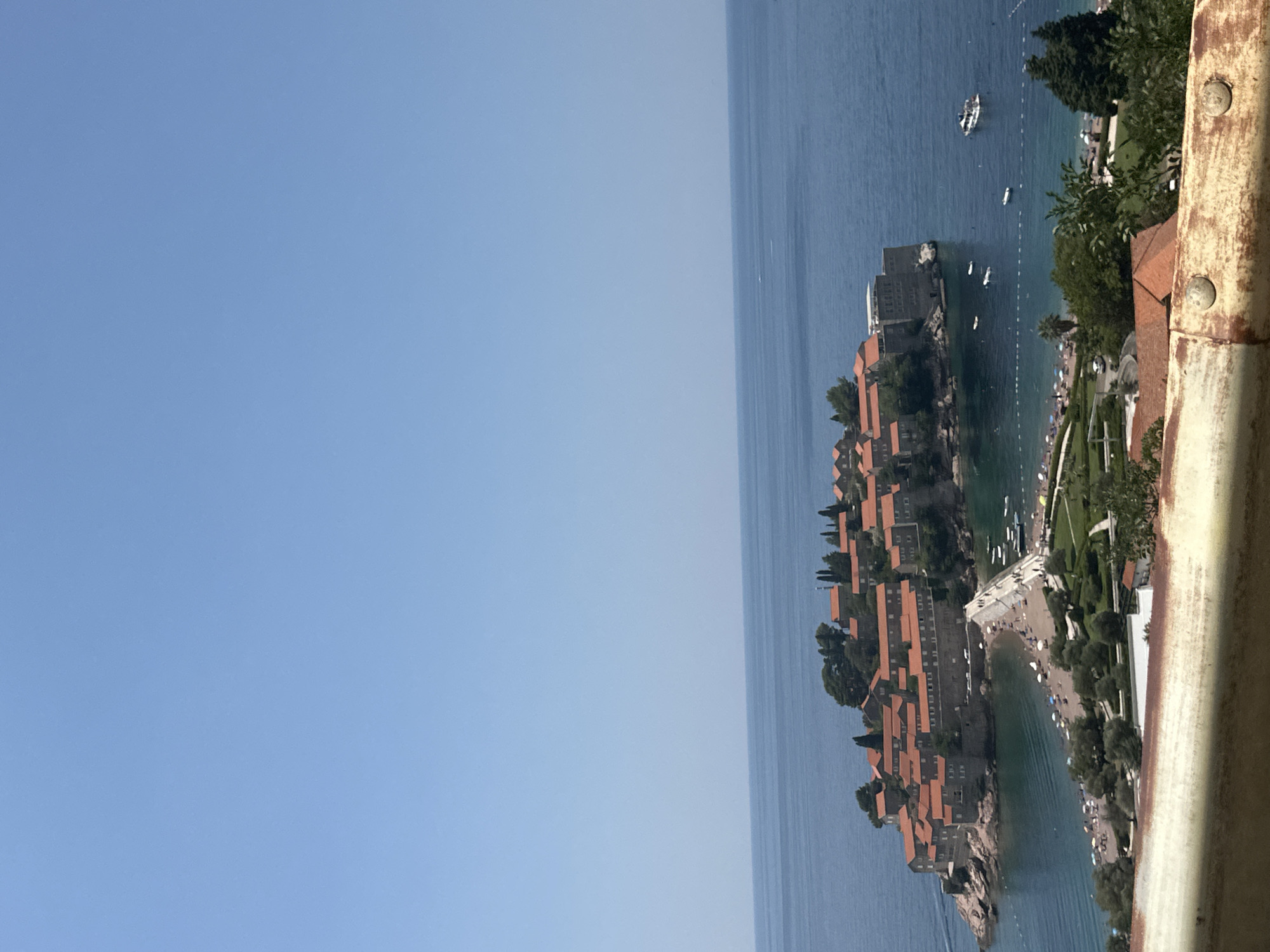 The image shows a panoramic view of Sveti Stefan, Montenegro, a fortified island village connected to the mainland by a narrow causeway. The island is densely packed with stone buildings featuring red-tiled roofs, nestled on a rocky outcrop. A causeway lined with people leads from the mainland to the island. The mainland features a beach with numerous umbrellas and sunbathers, as well as green landscaped areas with trees and some buildings. The sea surrounding the island is a clear blue, with several boats visible in the water. The sky is a clear, bright blue with no clouds. In the foreground, a weathered, rusty metal railing runs horizontally across the bottom of the frame.