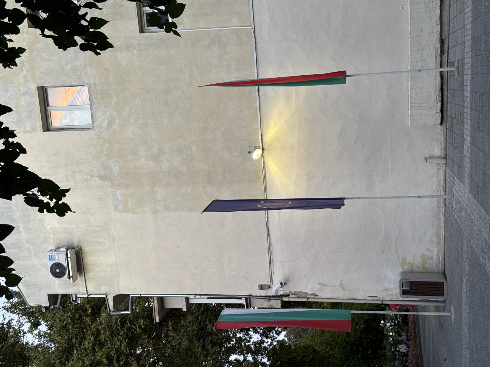 The image captures a section of a light-colored building exterior on a cloudy day, likely in the city of Plovdiv, Bulgaria. To the left, a large tree with dark green foliage partially obscures the upper left corner. Mounted on the building's wall is a white air conditioning unit. Below the AC unit, a security camera is visible, connected by wires running along the wall.
Three flagpoles stand vertically in front of the building. The leftmost flagpole displays a Bulgarian flag, with its horizontal stripes of white, green, and red visible. Next to it, a flagpole displays a European Union flag, a deep blue rectangle with a circle of yellow stars. The rightmost flagpole shows a flag with vertical red and green stripes.
At the base of the building, a grey trash receptacle with wooden slats is situated. The ground in front is paved with grey and white patterned tiles. In the background, a blurry scene depicts a park-like area with people seated and standing, and a pink-clad figure partially visible. A window on the building's second floor reflects the sky, showing orange and white hues. A light fixture on the exterior wall casts a warm glow downwards. The overall impression is a quiet urban scene, possibly in a public square or walkway.