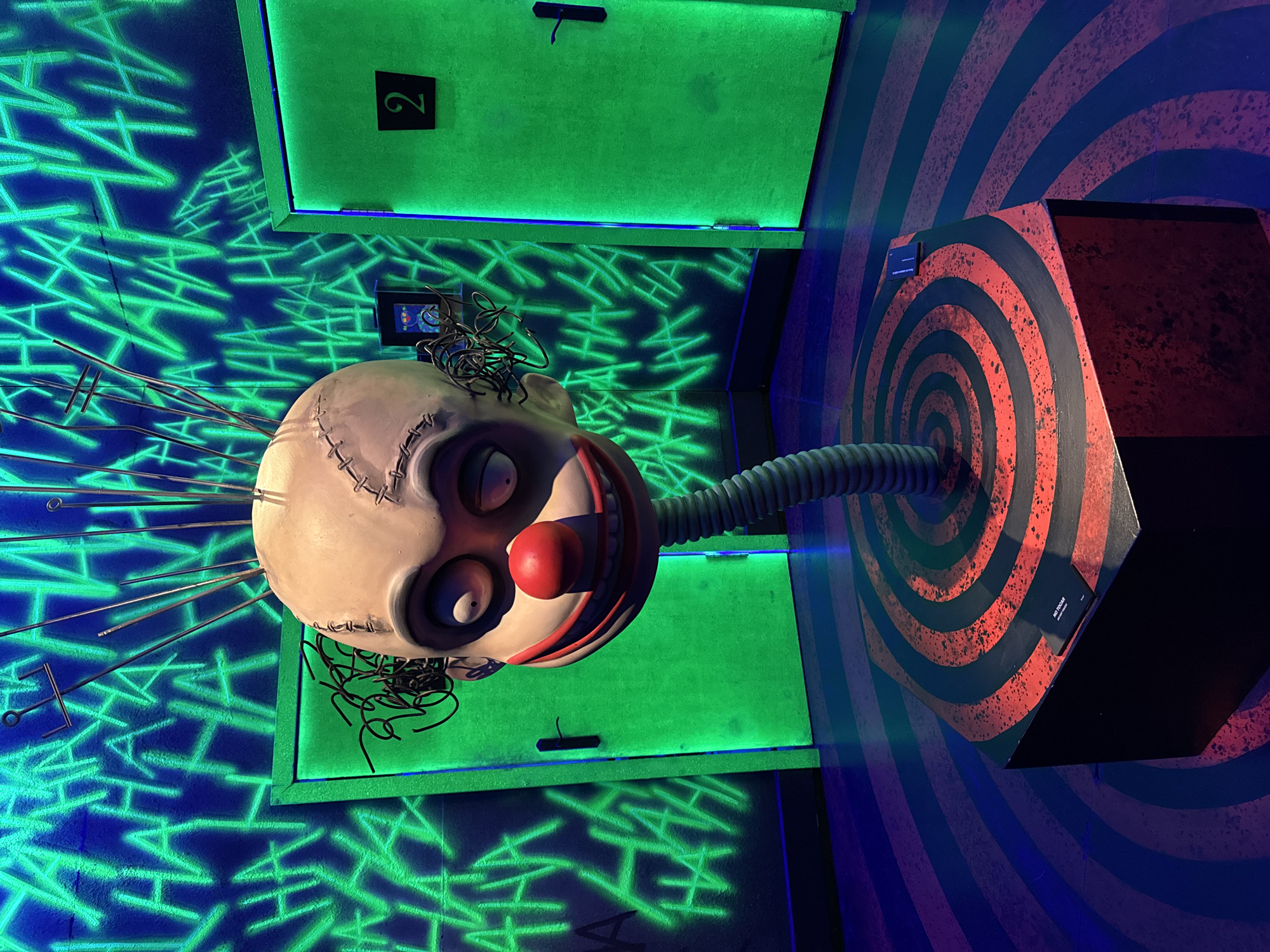 In a darkly lit room with vibrant neon green lighting, a bizarre sculpture of a clown's head is prominently displayed. The clown's head is pale and scarred, with large, discolored eyes and a bright red nose. It is attached to a flexible, corrugated neck and stands atop a hexagonal pedestal with a red and black spiral design. The background features walls covered in neon green "HAHA" graffiti, and two identical green doors, one with the number "2" on it. The floor has a swirling pattern of dark blue and red stripes. There are no people visible in the image. The overall atmosphere is unsettling and theatrical, suggesting a theme park or entertainment venue. This sculpture is located at Cintermex in Monterrey, Mexico.